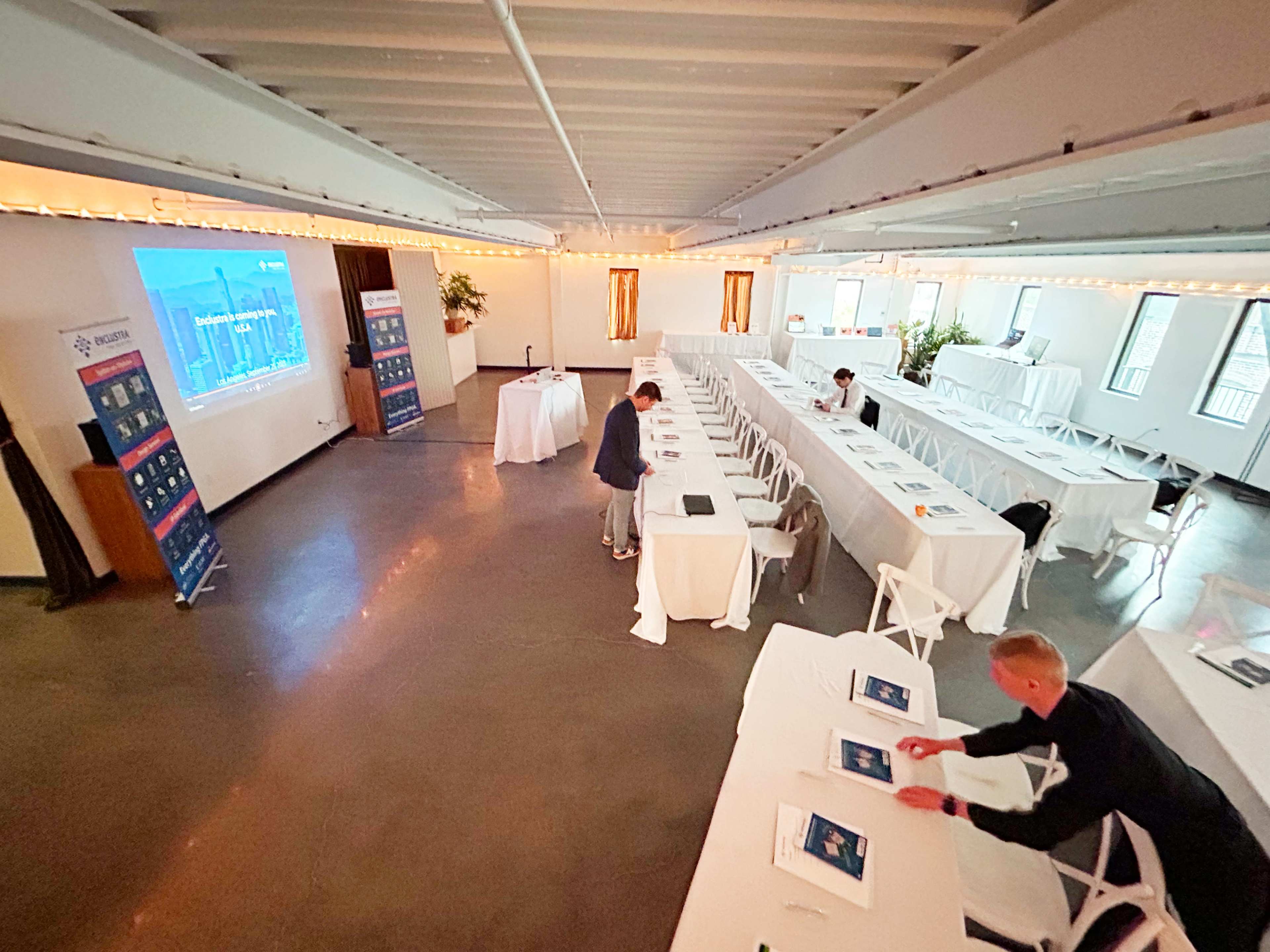 A conference room is set up with long white tables, chairs, and a presentation screen, while two individuals prepare materials before an event.