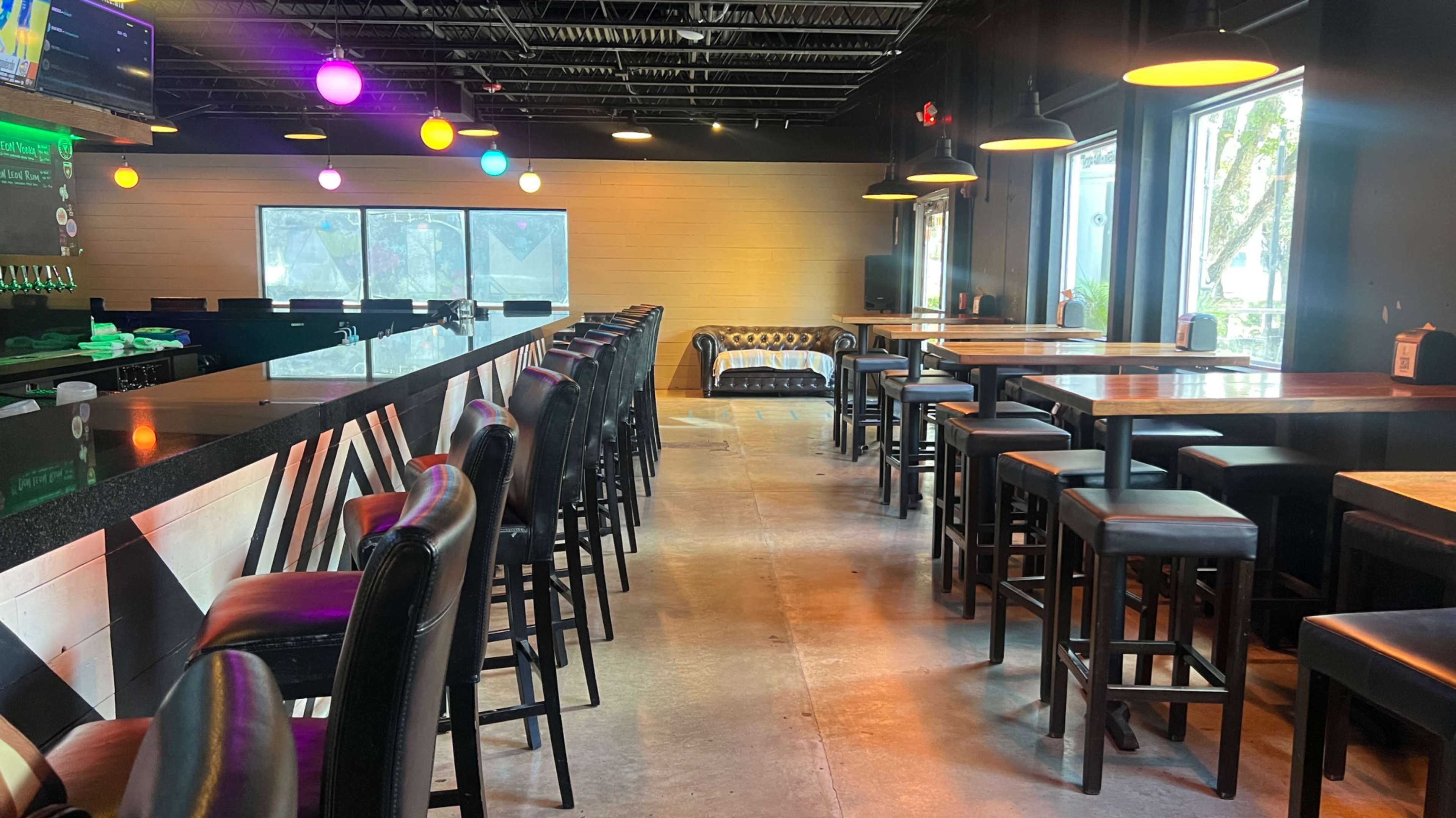 The image shows the interior of a modern bar with a long counter, high stools, and a few tables, illuminated by colorful pendant lights.