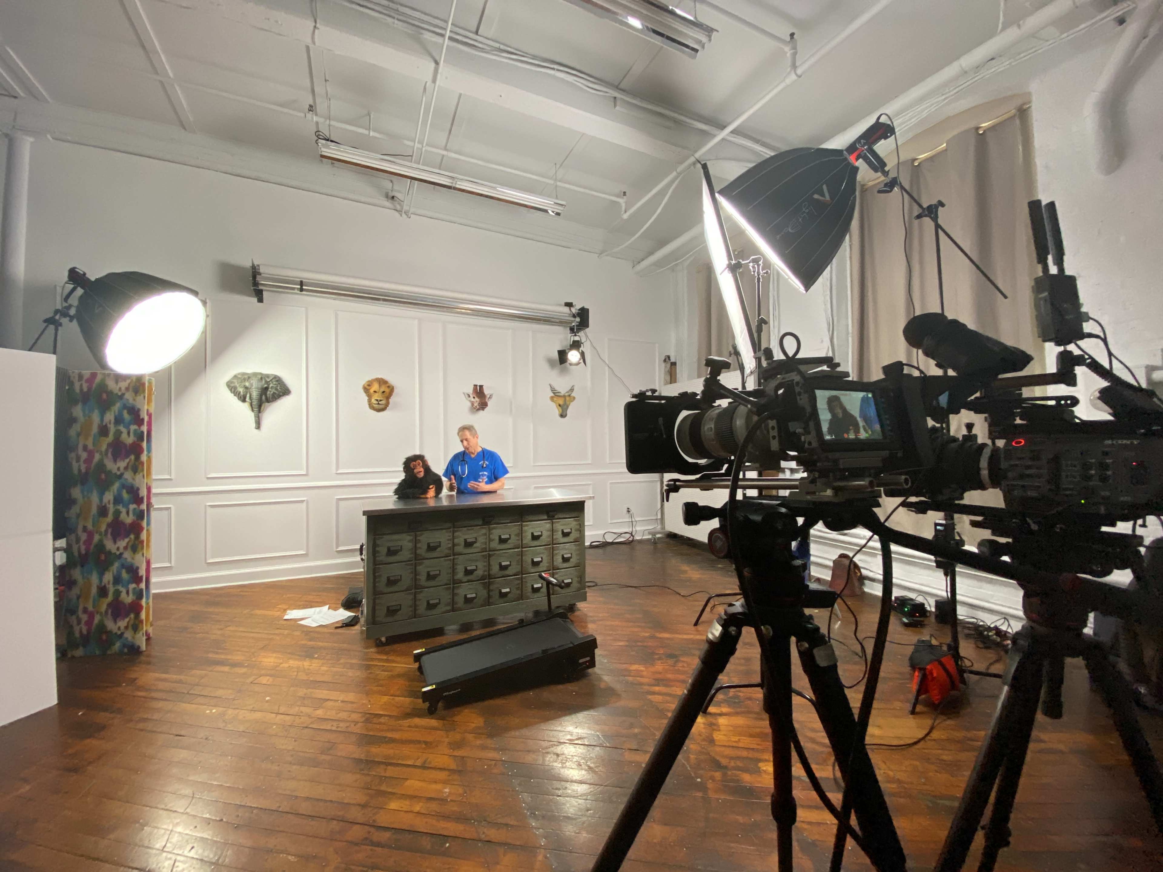 A film crew sets up equipment in a studio featuring a large wooden desk and various lighting setups.