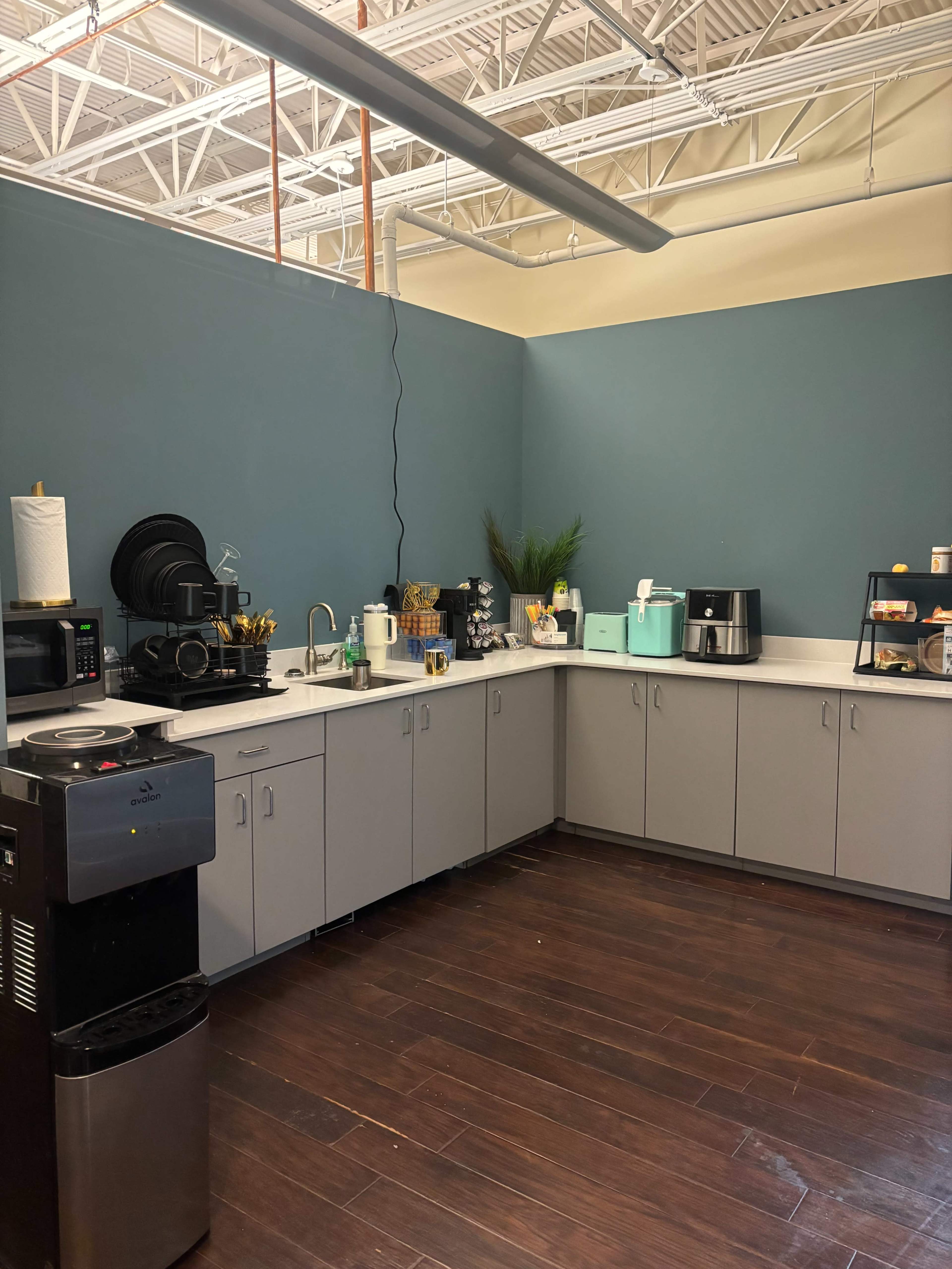 The image shows a modern kitchen area with gray cabinets, a coffee station, and various appliances against a blue wall.