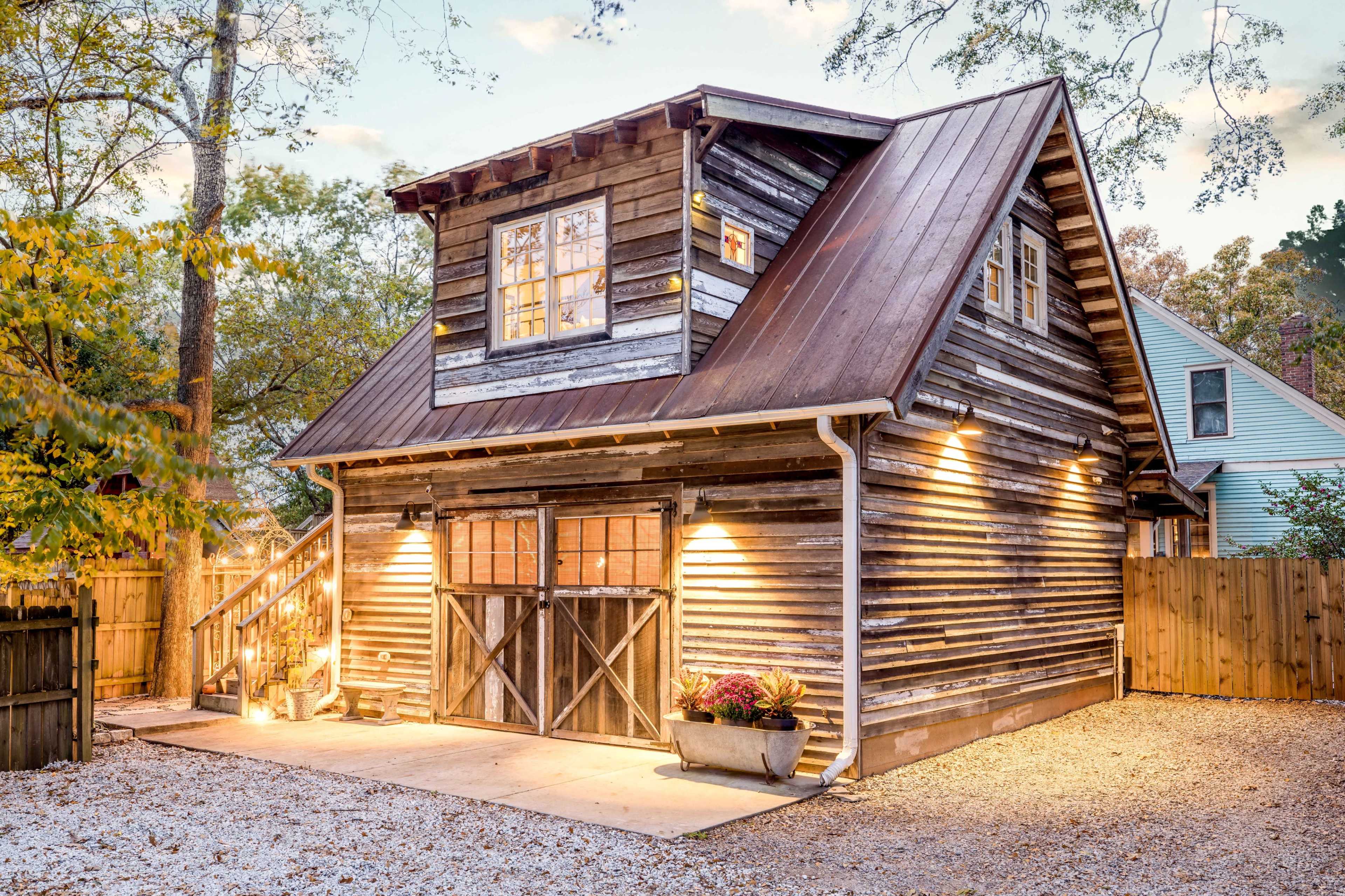 A rustic wooden house with a metal roof, illuminated by soft outdoor lighting, surrounded by trees and a wooden fence.