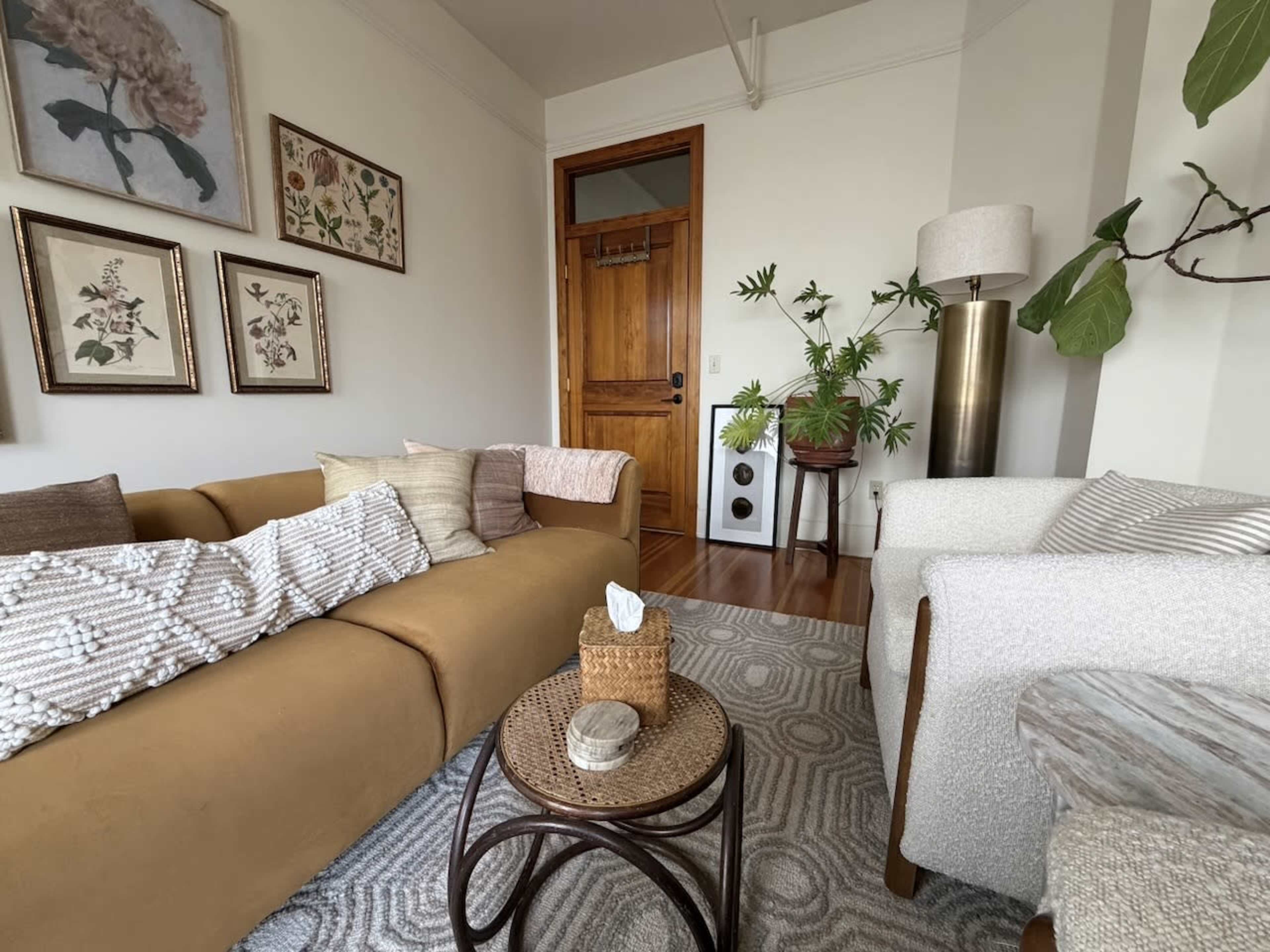 The image shows a cozy living room with a brown sofa, a white armchair, decorative wall art, a wooden door, and a potted plant.