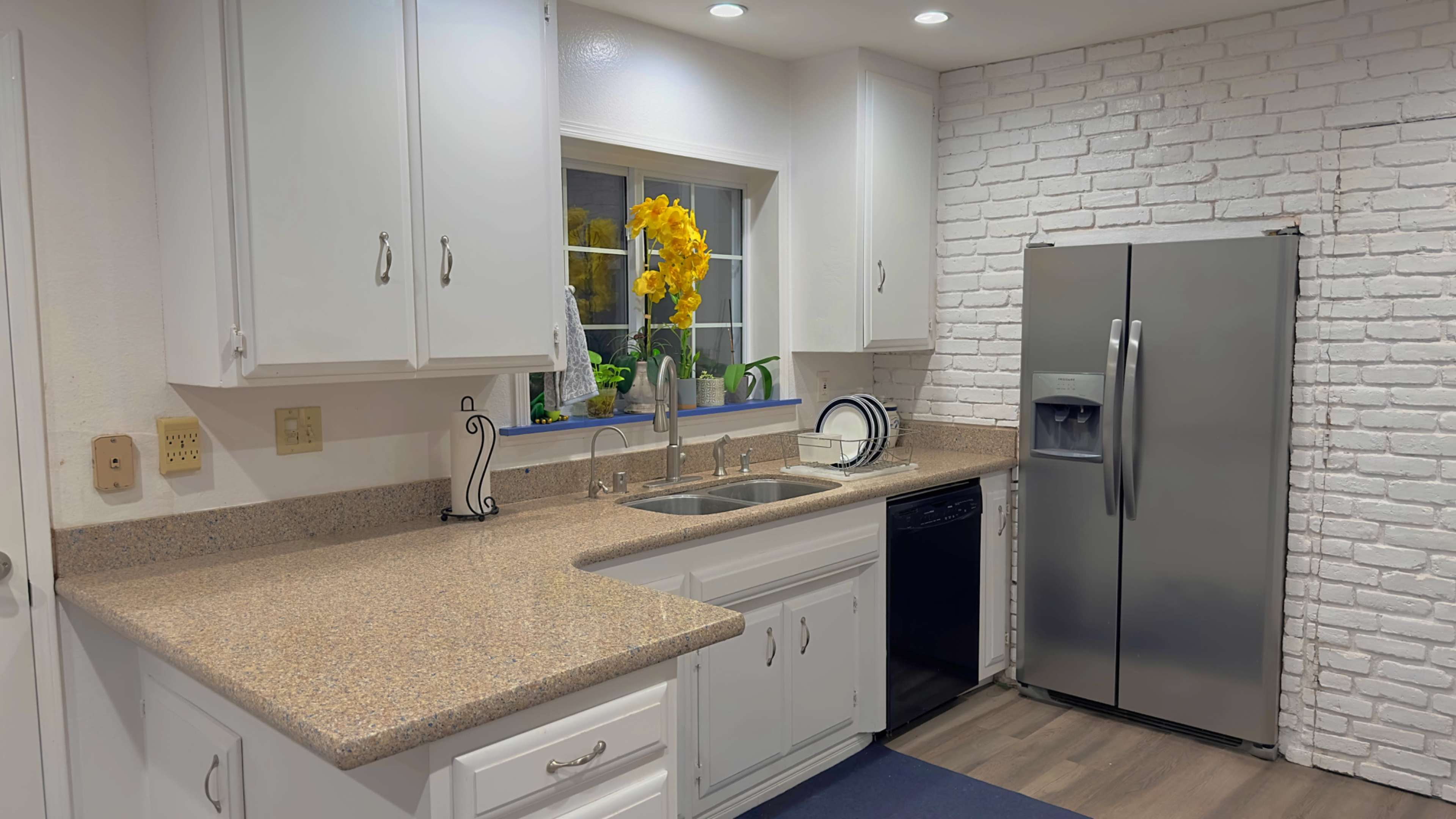 A clean kitchen with white cabinetry, a stainless steel refrigerator, and a window with a vase of yellow flowers on the counter.