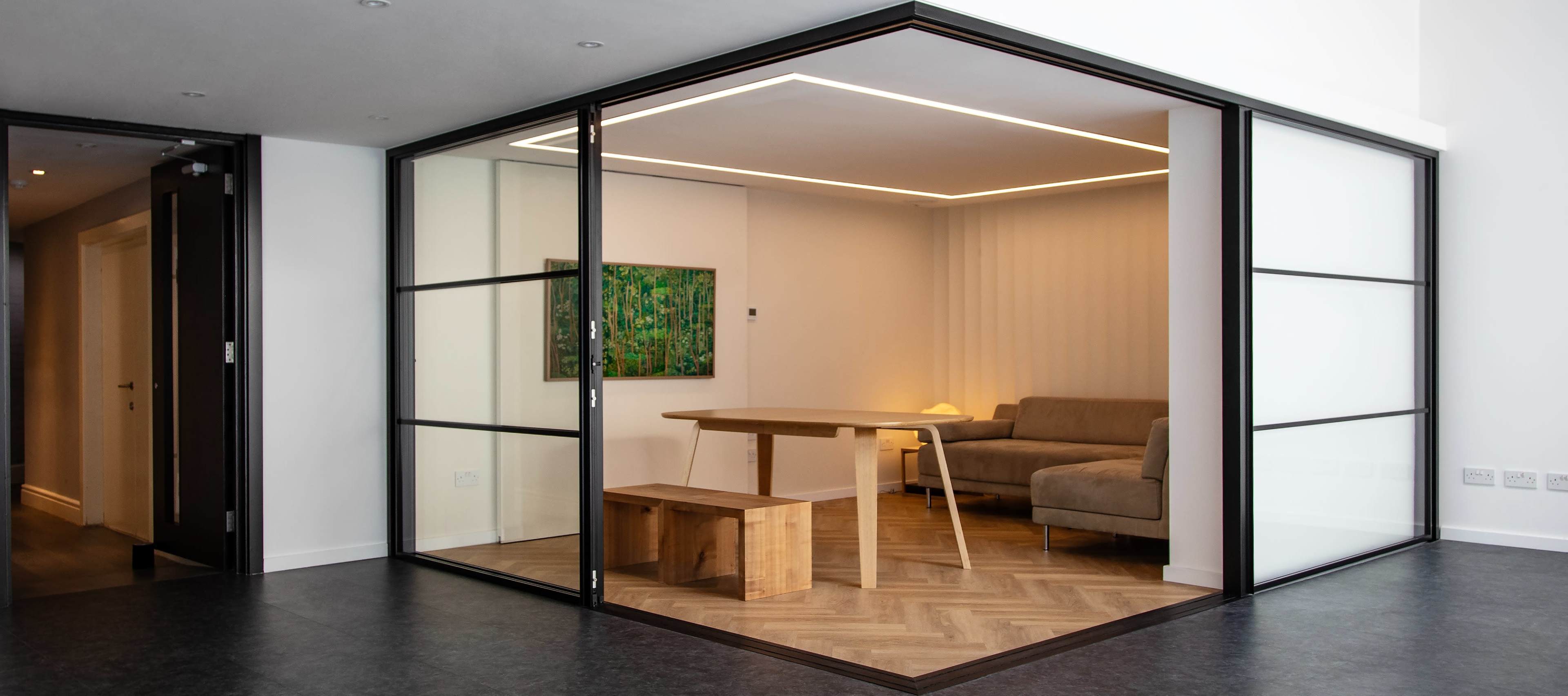 The image shows a modern glass-enclosed room featuring a sofa, a wooden table, and a bench against a backdrop of minimalistic decor.