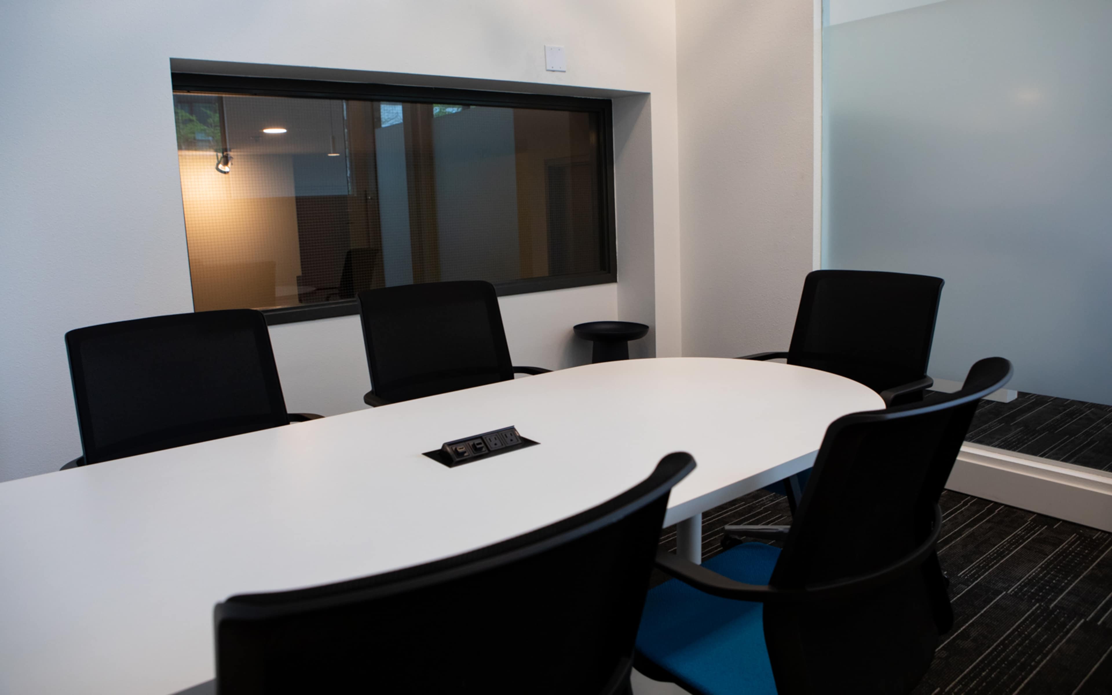 A modern conference room features a white oval table surrounded by black chairs, with a window allowing natural light and a wall-mounted power outlet on the table.