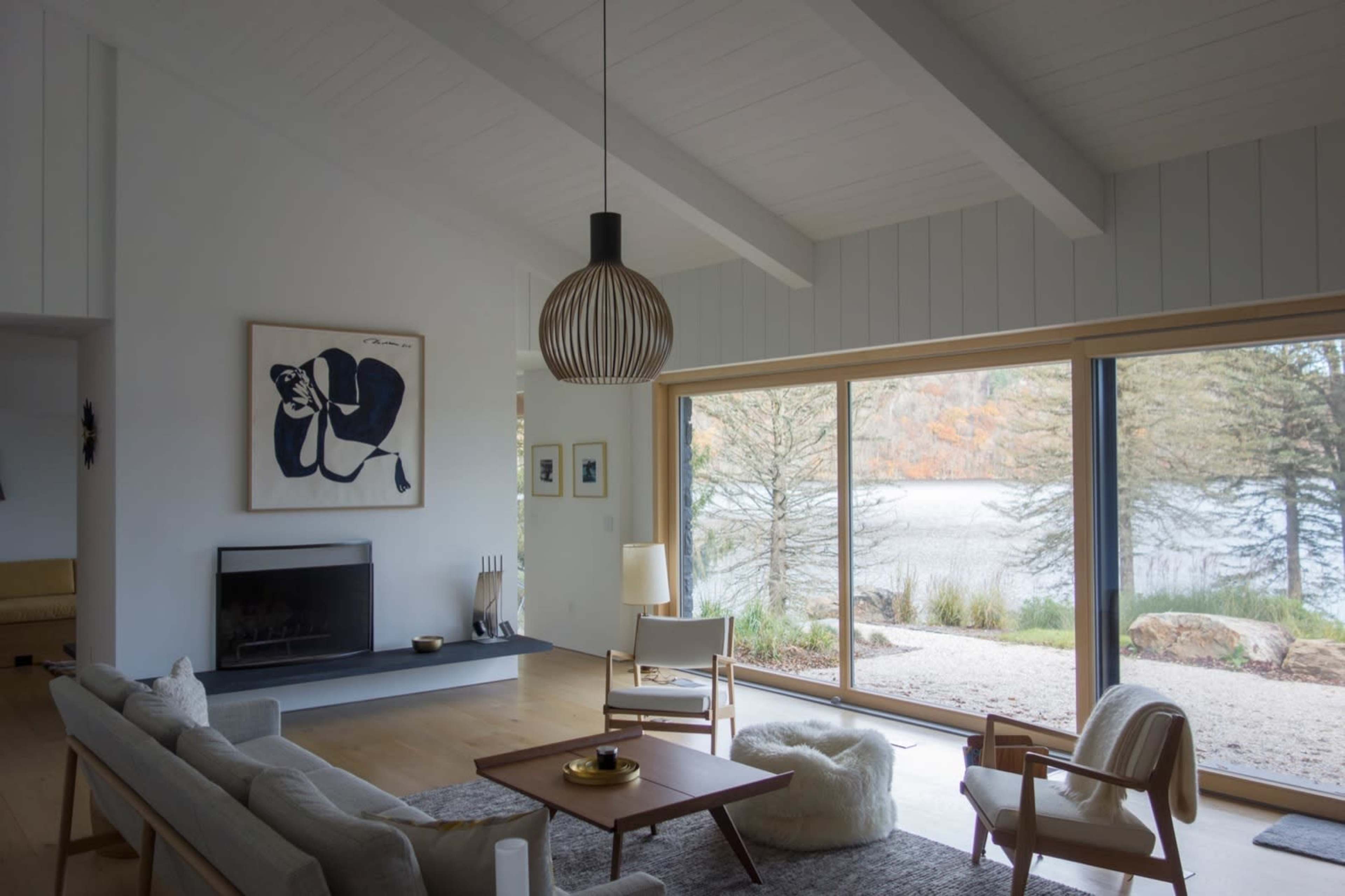 The image shows a modern living room with a large window overlooking a lake, featuring minimalist furniture and a simple, elegant design.