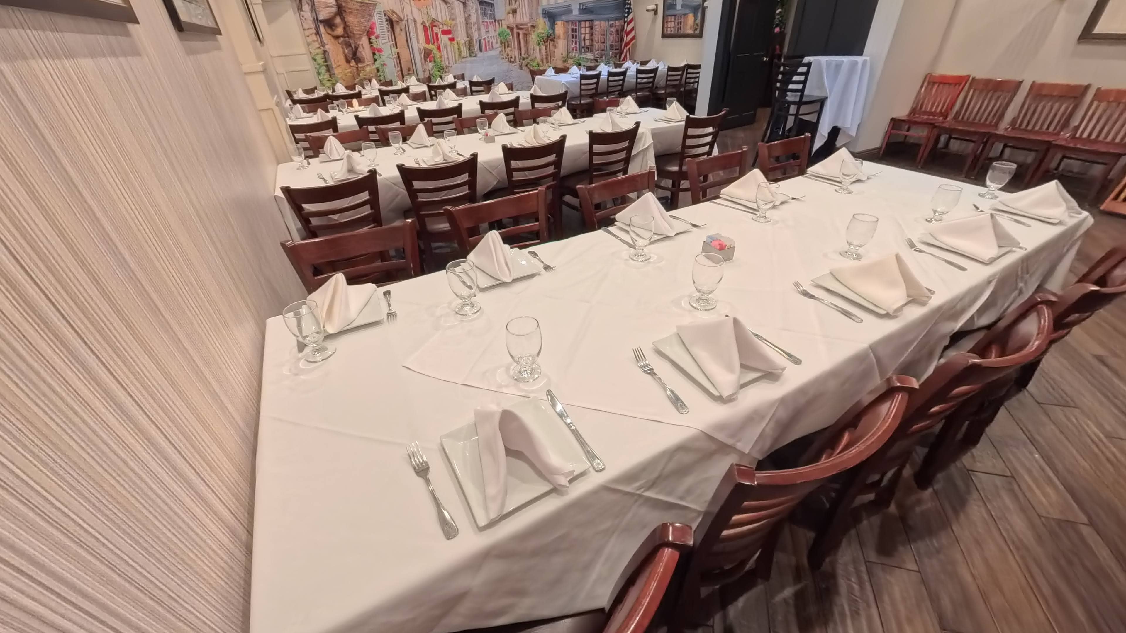 A neatly set dining table with white tablecloths and folded napkins is arranged in a restaurant, surrounded by rows of wooden chairs.