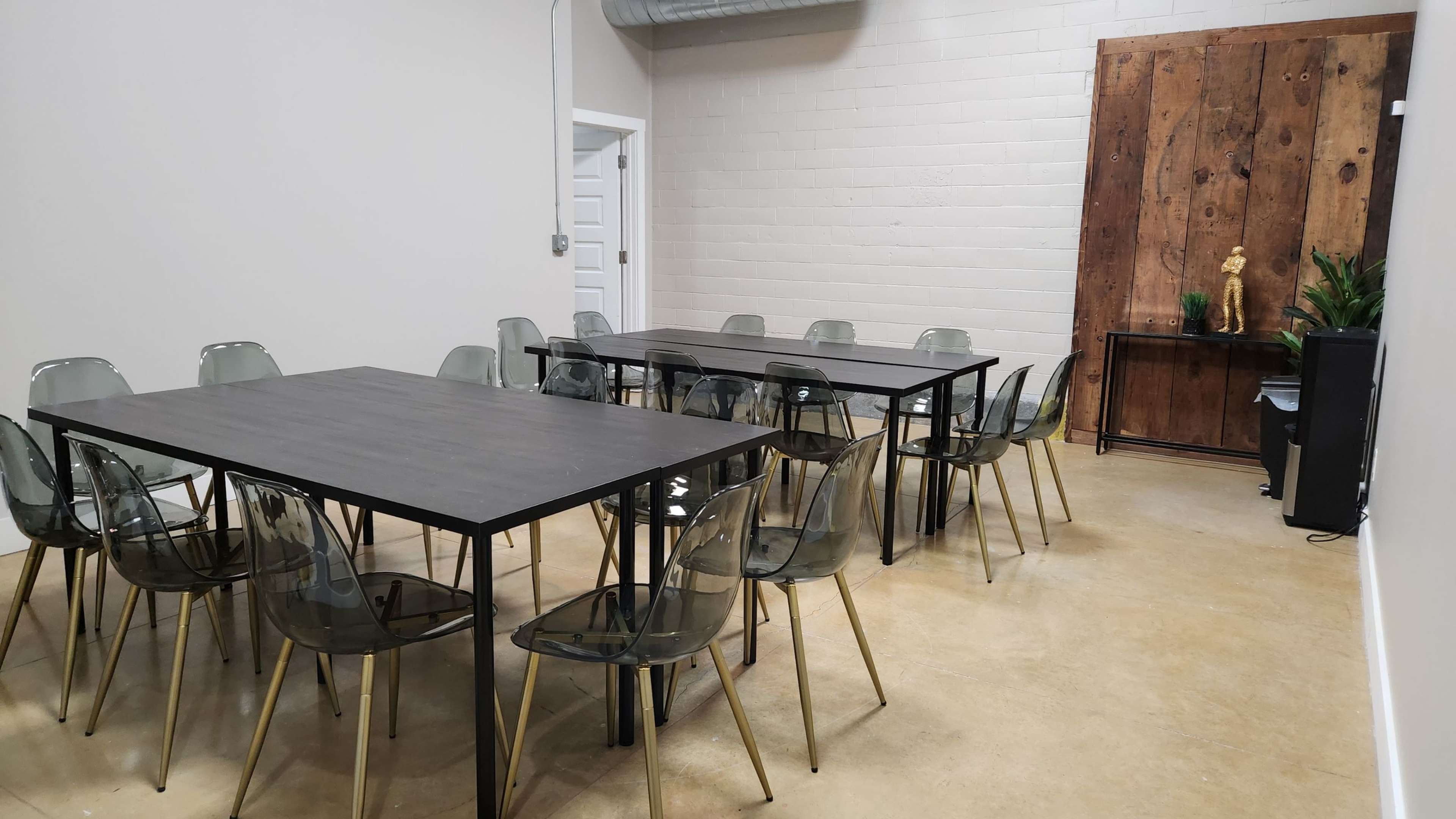 A modern meeting room features two rectangular black tables surrounded by clear acrylic chairs, with a wooden accent wall and a small shelf displaying decorative items.