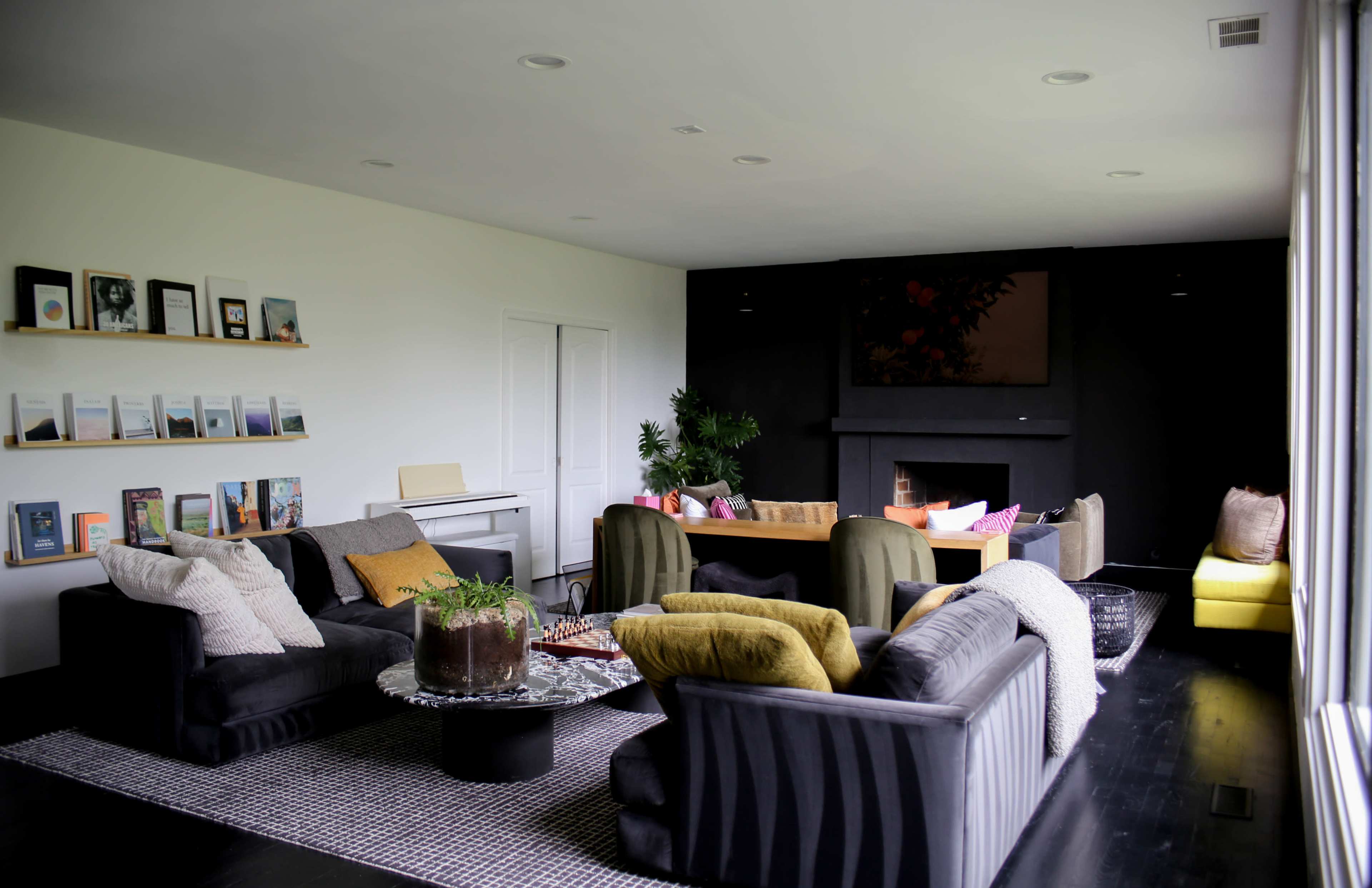 The image shows a modern living room with several sofas arranged around a round coffee table, a bookshelf on the wall, and large windows letting in natural light.