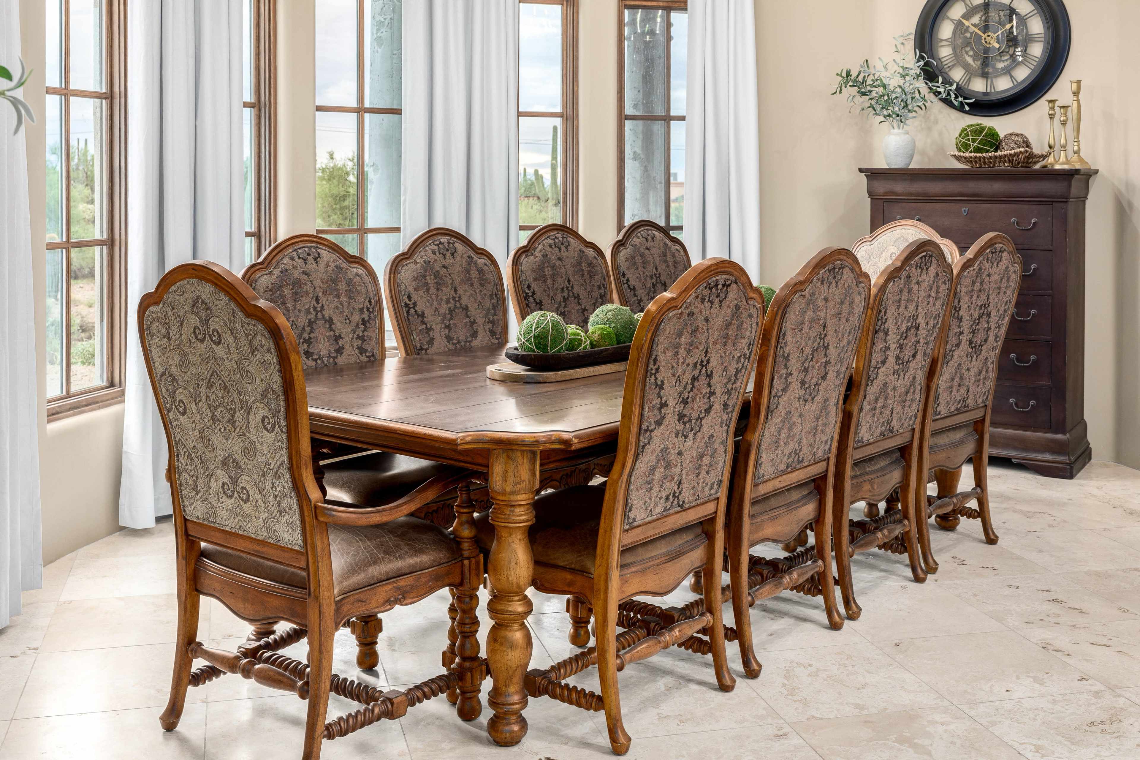 A long wooden dining table is surrounded by eight upholstered chairs, set near large windows with white curtains and a decorative cabinet in the background.