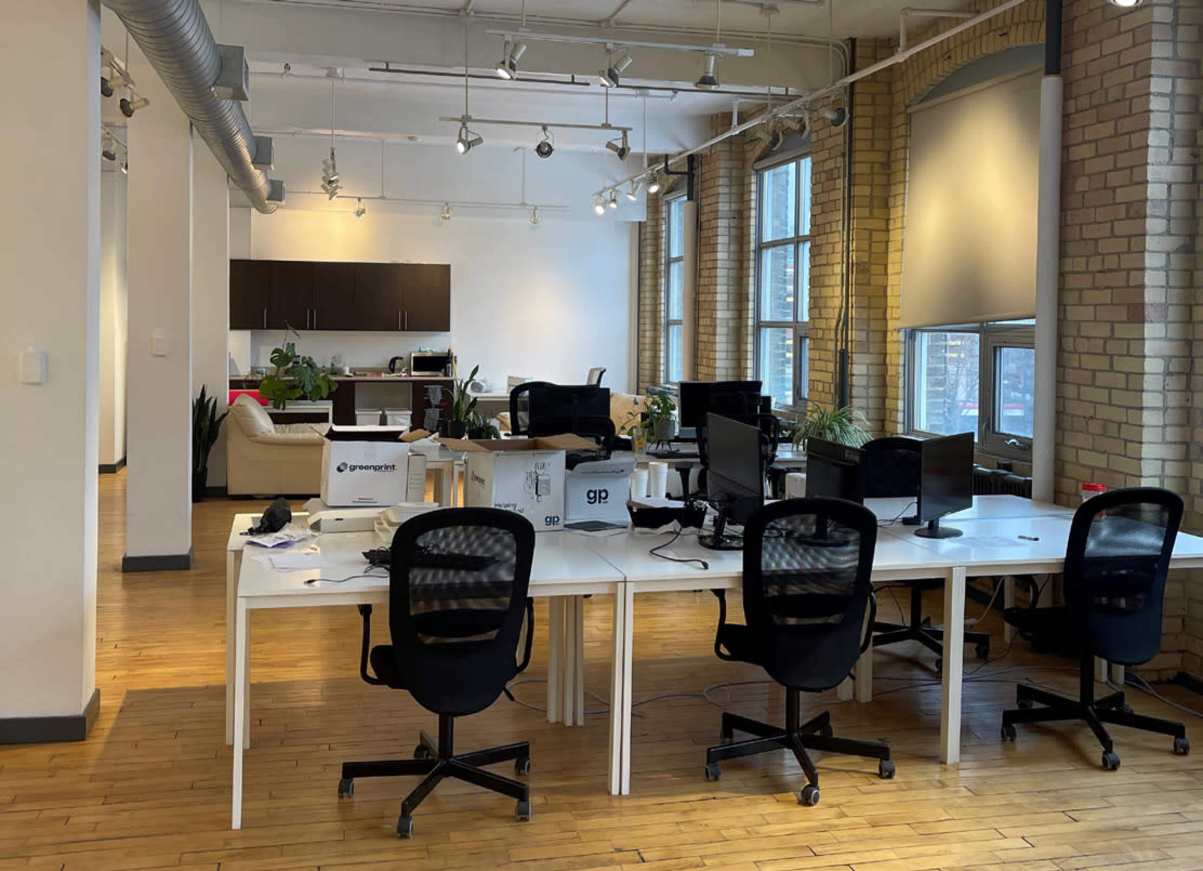 A spacious office with several desks, computer chairs, and large windows, featuring a lounge area in the background.
