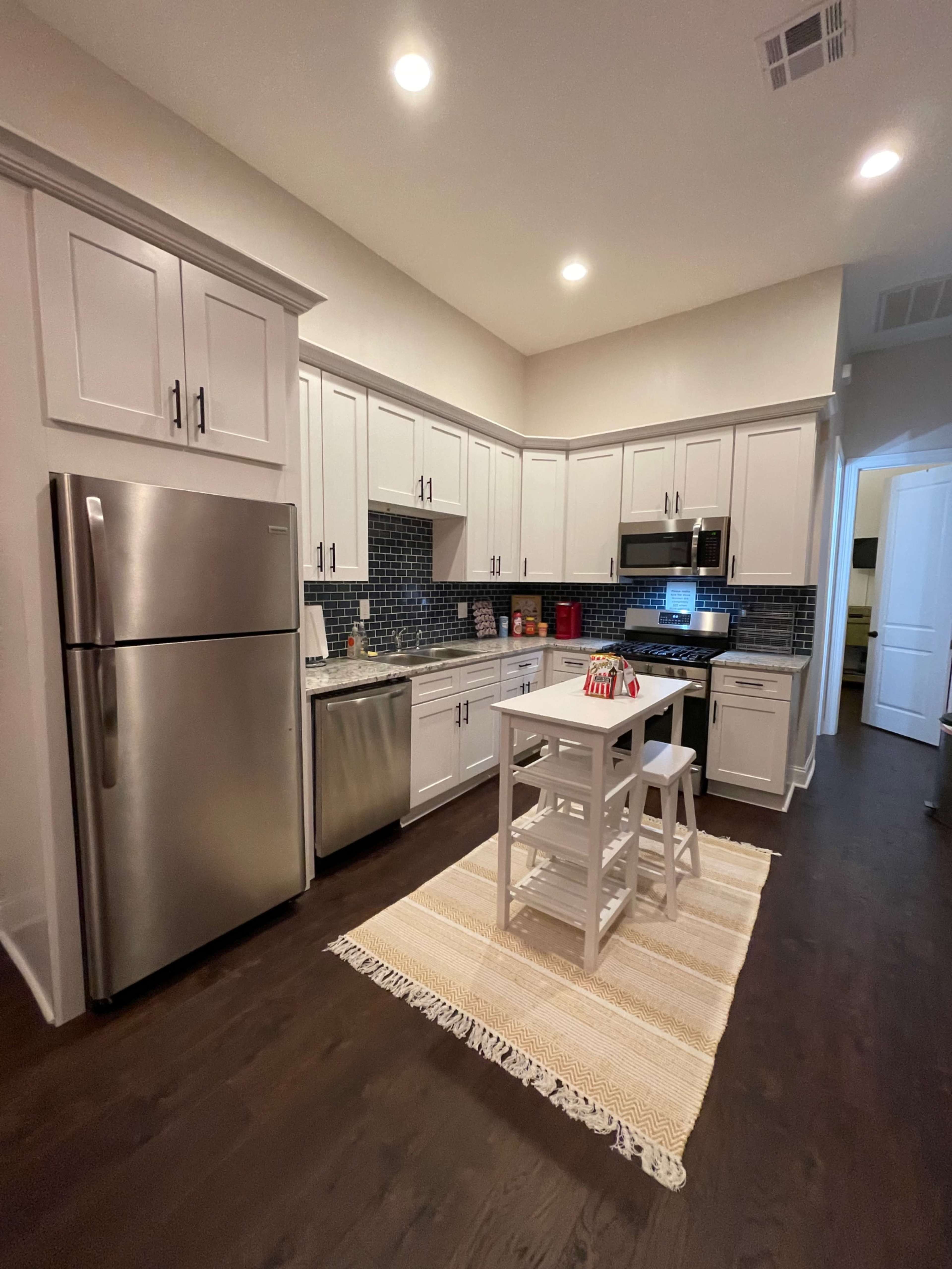 The image shows a modern kitchen with stainless steel appliances, white cabinetry, and a small dining table set in the center.