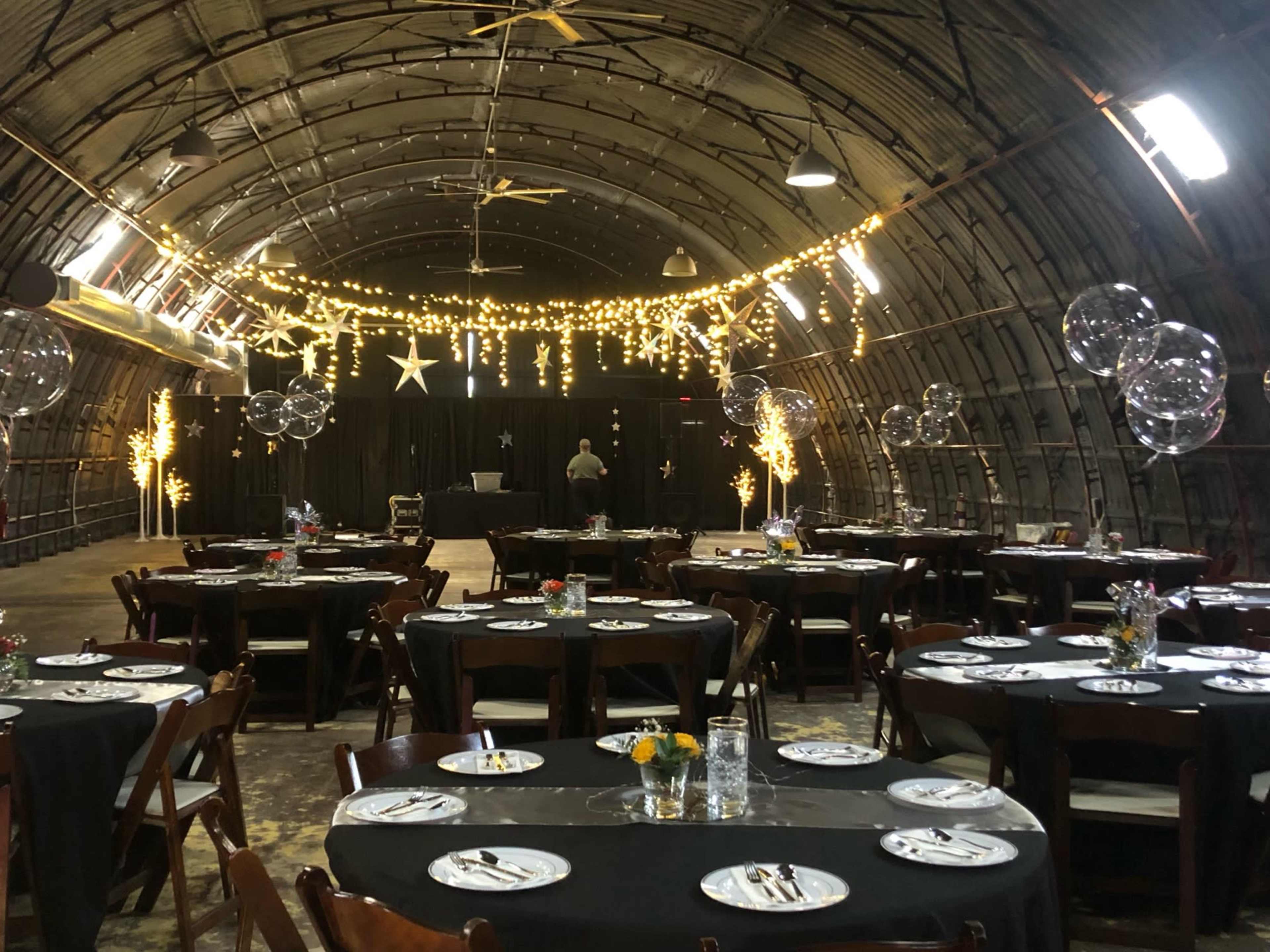 The image shows an elegantly arranged dining area inside a large, curved space, with tables set up for an event and decorative lights hanging from the ceiling.