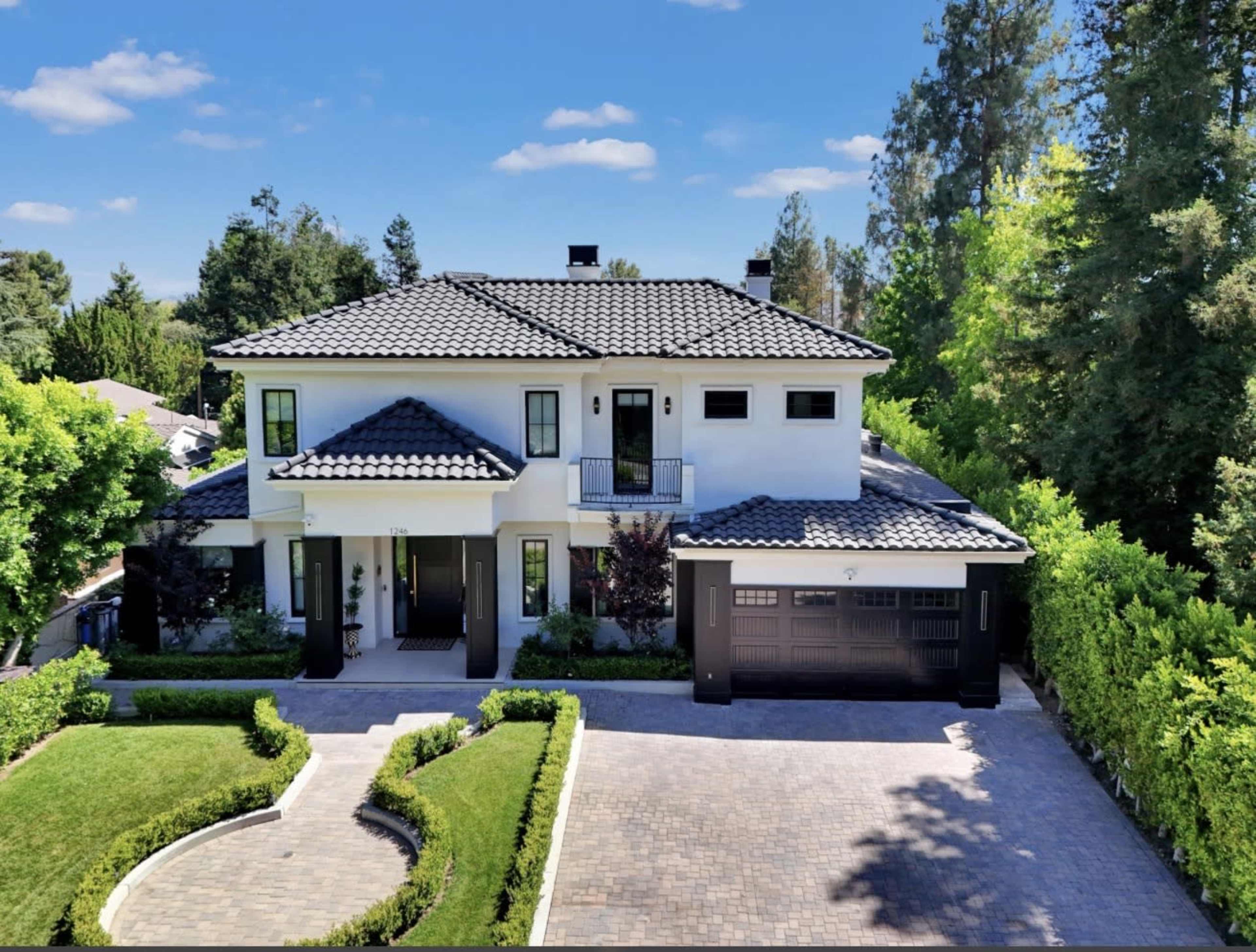 The image shows a modern, two-story white house with a tile roof, surrounded by greenery and a cobblestone driveway.