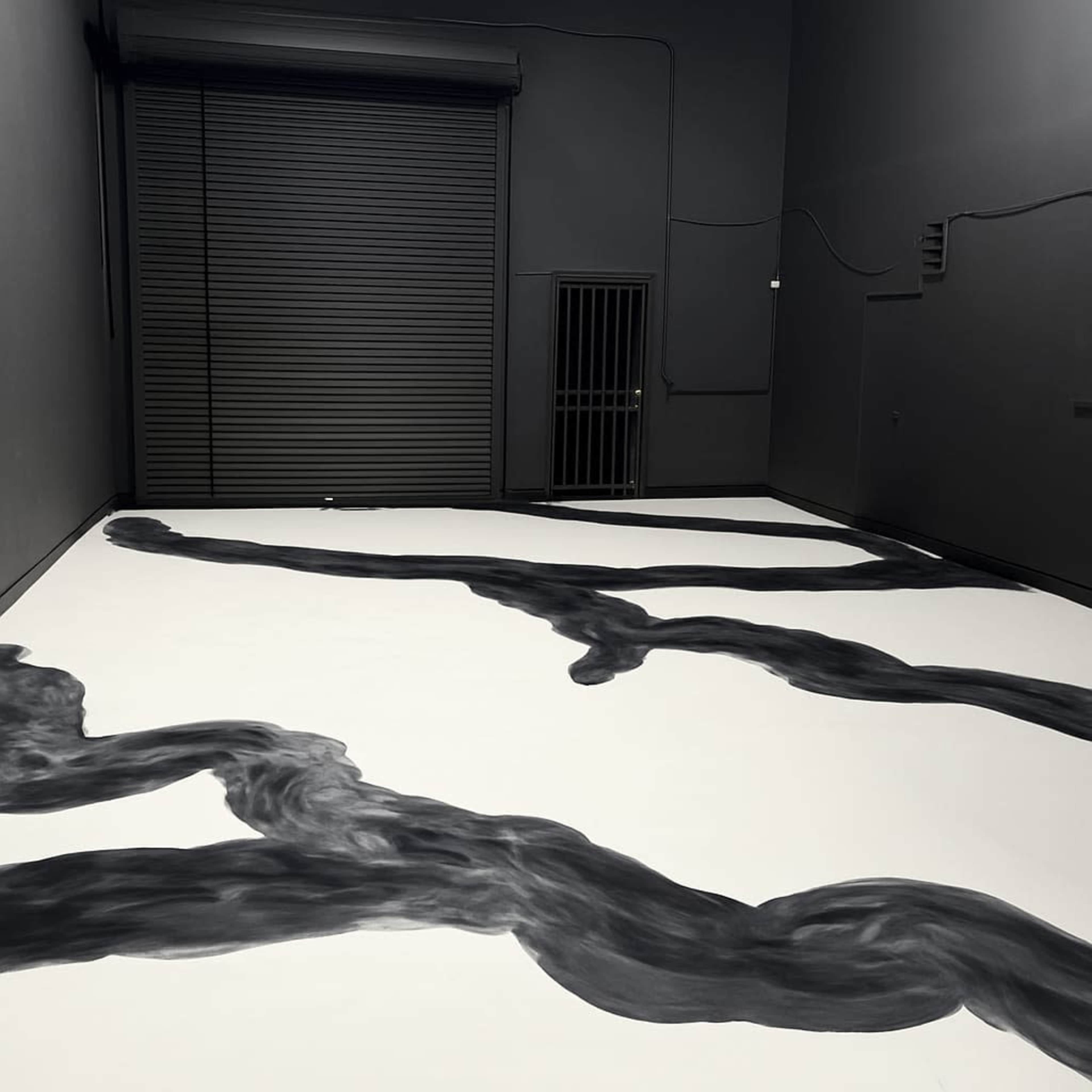 The image shows a dimly lit room with a gray floor that features a large, abstract black painting resembling flowing lines, and a closed metal door and barred window on one wall.