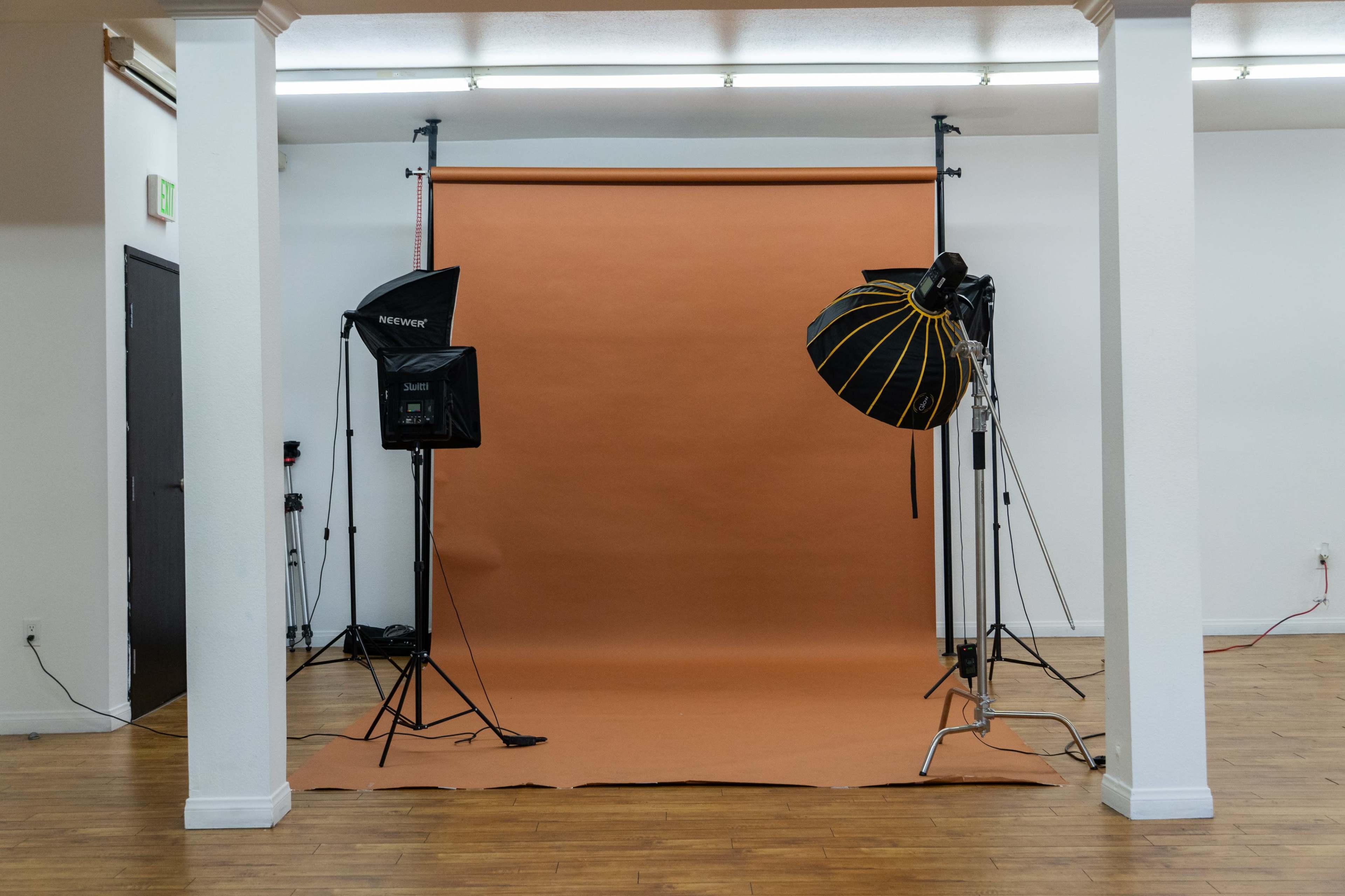 The image shows a photography studio with a brown backdrop, two studio lights on stands, and wooden flooring.