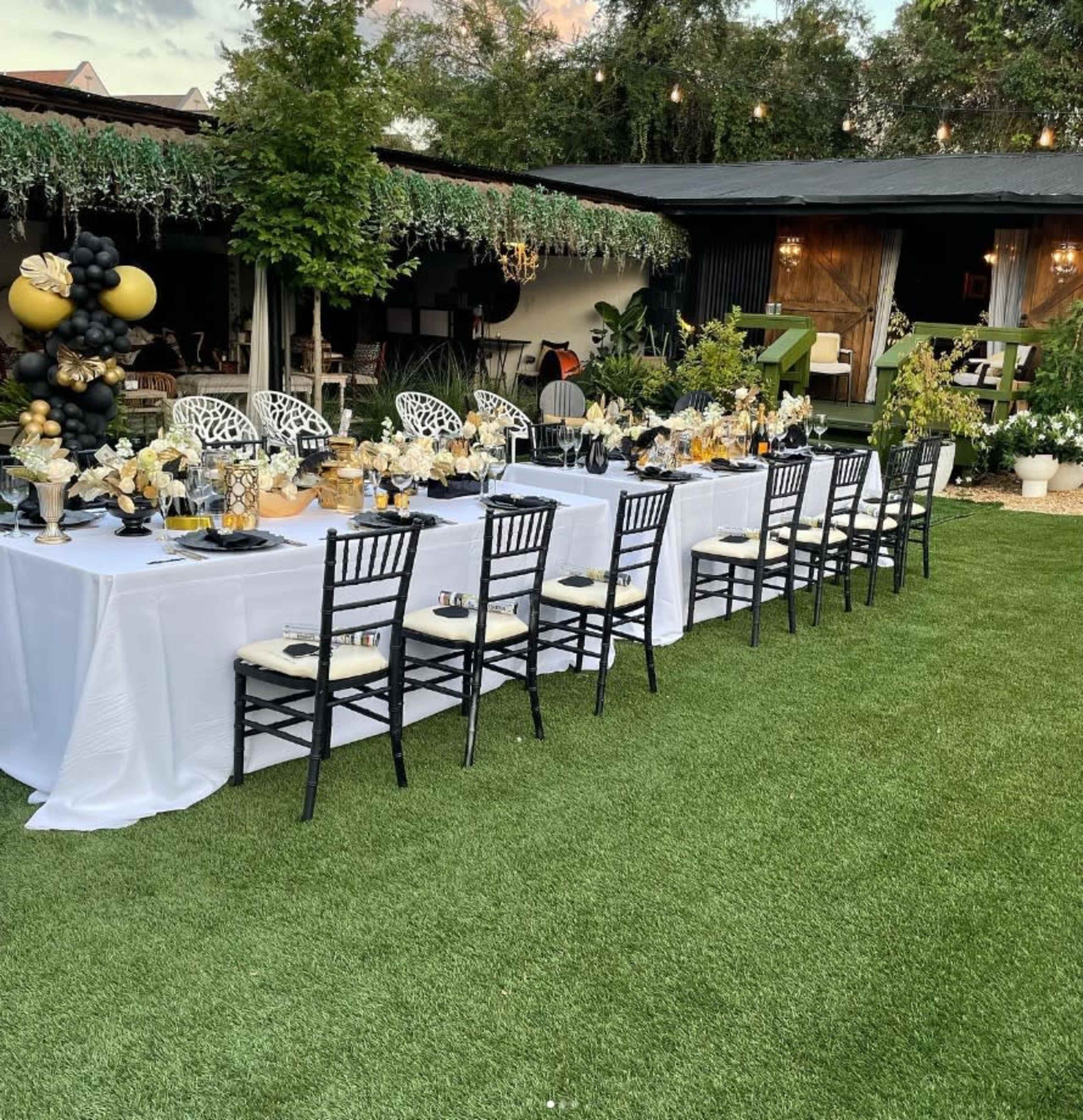 A long banquet table is set outdoors with elegant decor, surrounded by greenery and string lights.