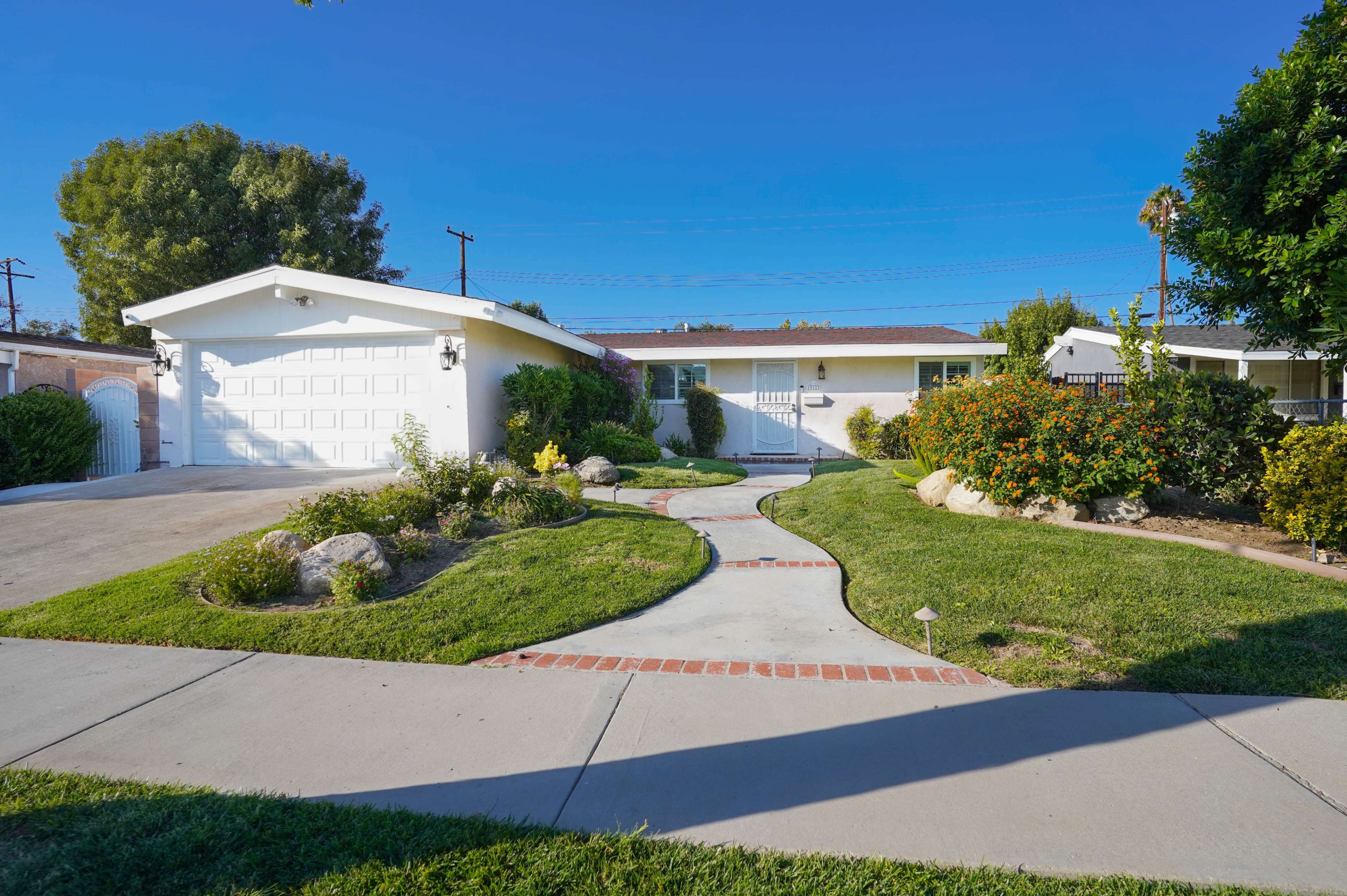 A single-story house with a garage is surrounded by a landscaped yard featuring a curved pathway and various plants.