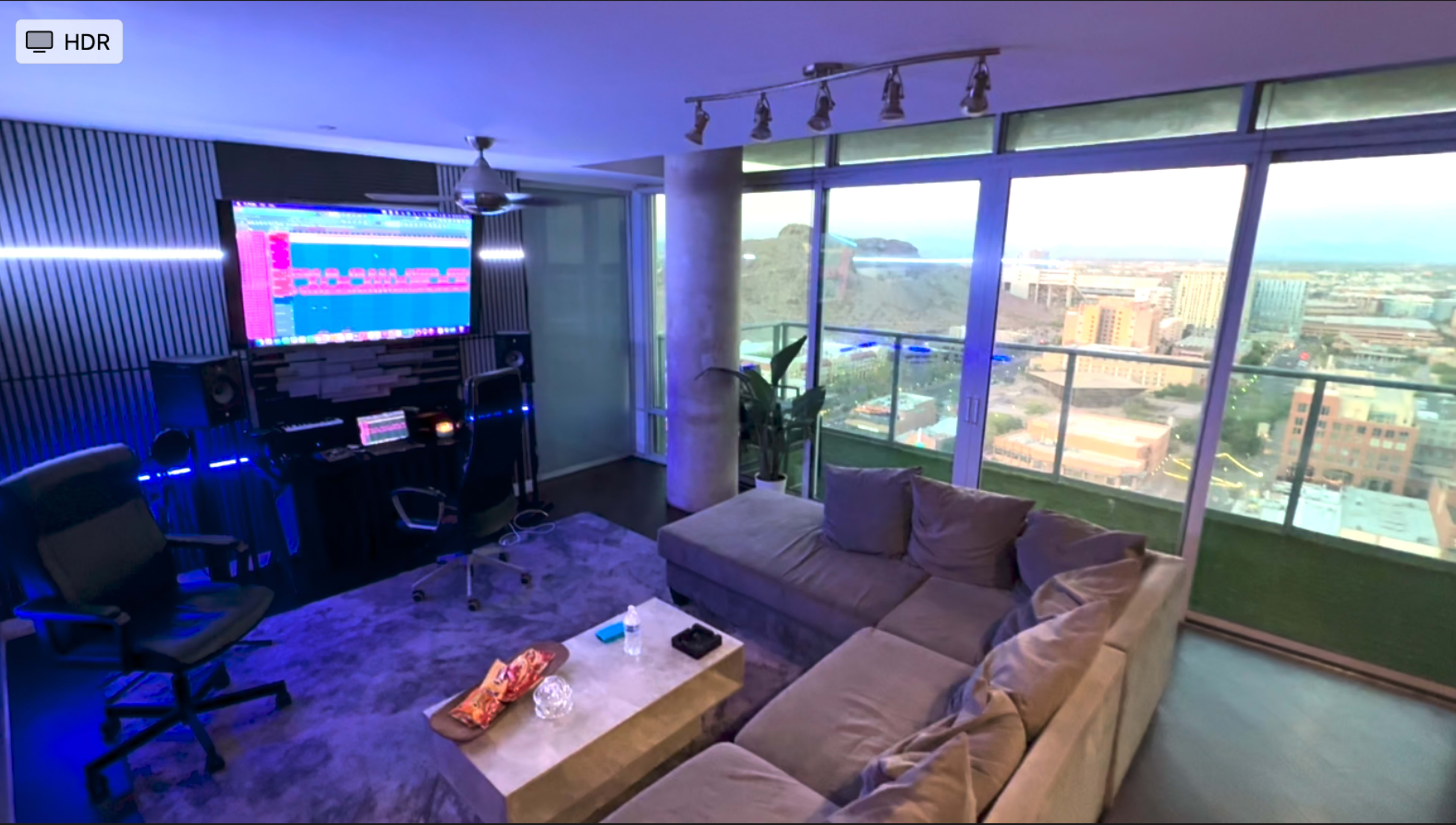 The image shows a modern living room with a large sectional sofa, a workspace featuring a computer setup against a window, and a cityscape view visible outside.