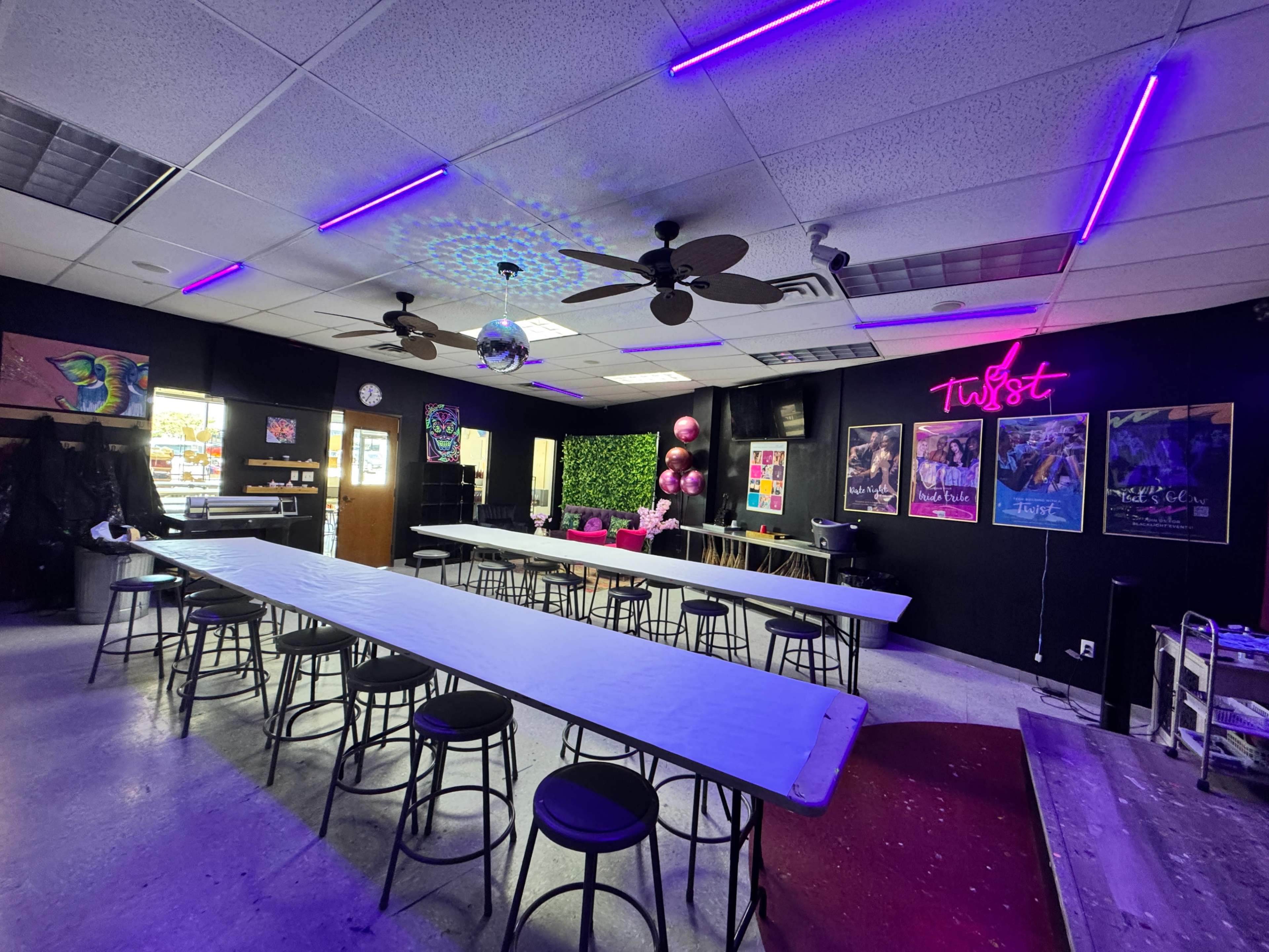 The image shows an indoor event space with multiple long tables, black stools, and neon lights, featuring decorative elements like disco balls and a green plant wall.