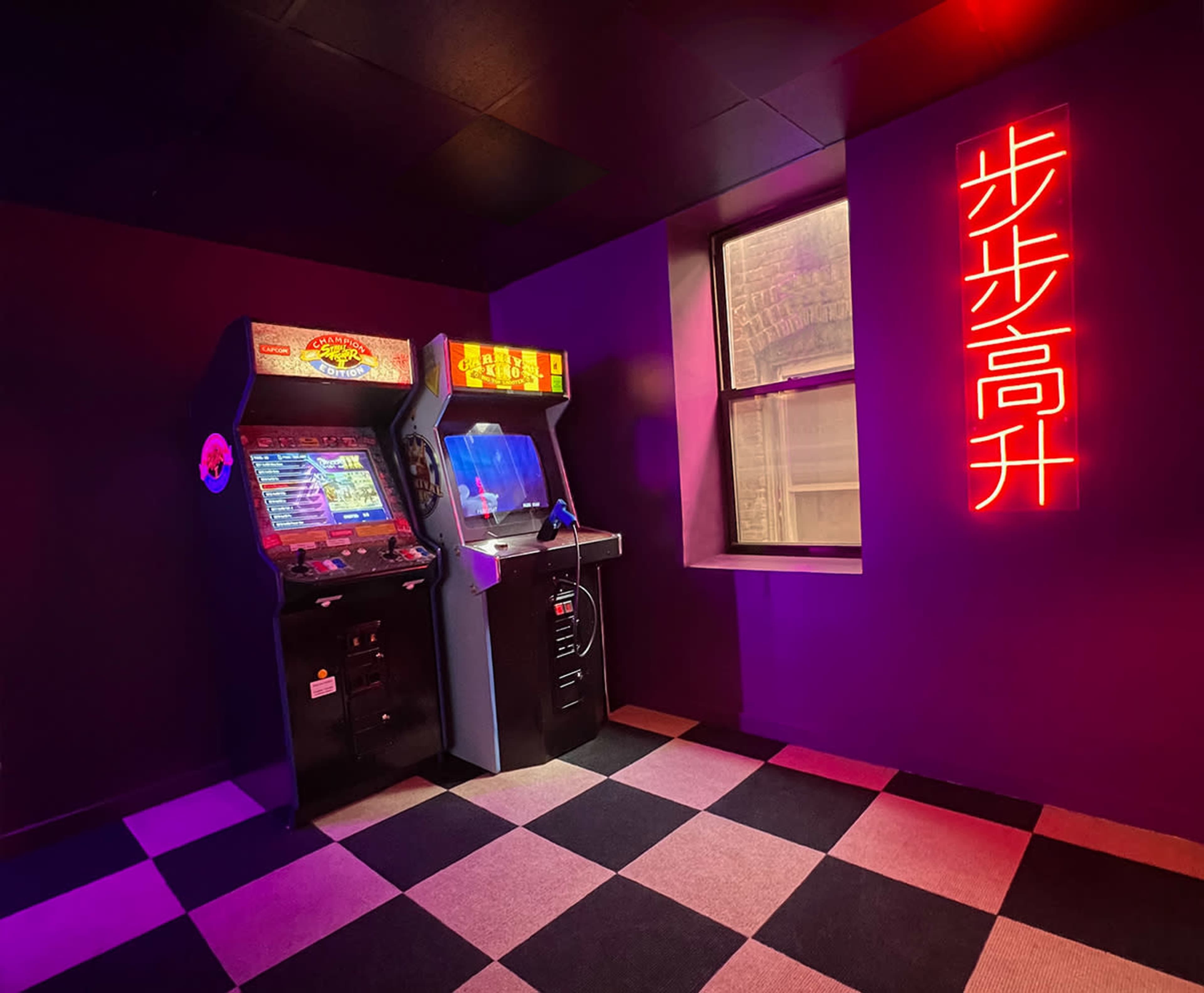 The image shows a dimly lit arcade room featuring a colorful arcade cabinet beside a monitor setup, with a neon sign on the wall.