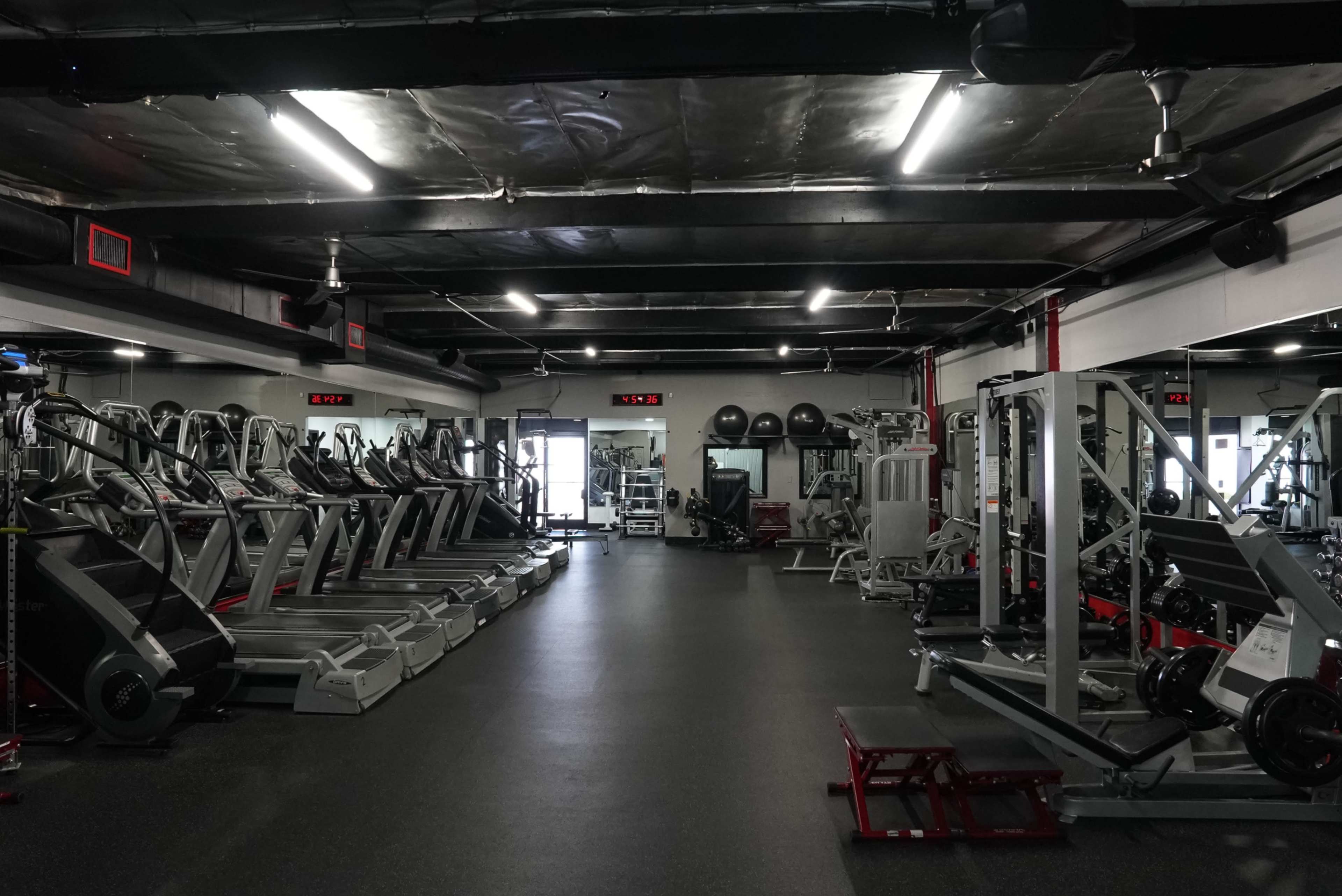 A modern gym interior with various exercise machines arranged in rows, including treadmills and weight machines, illuminated by overhead lights.