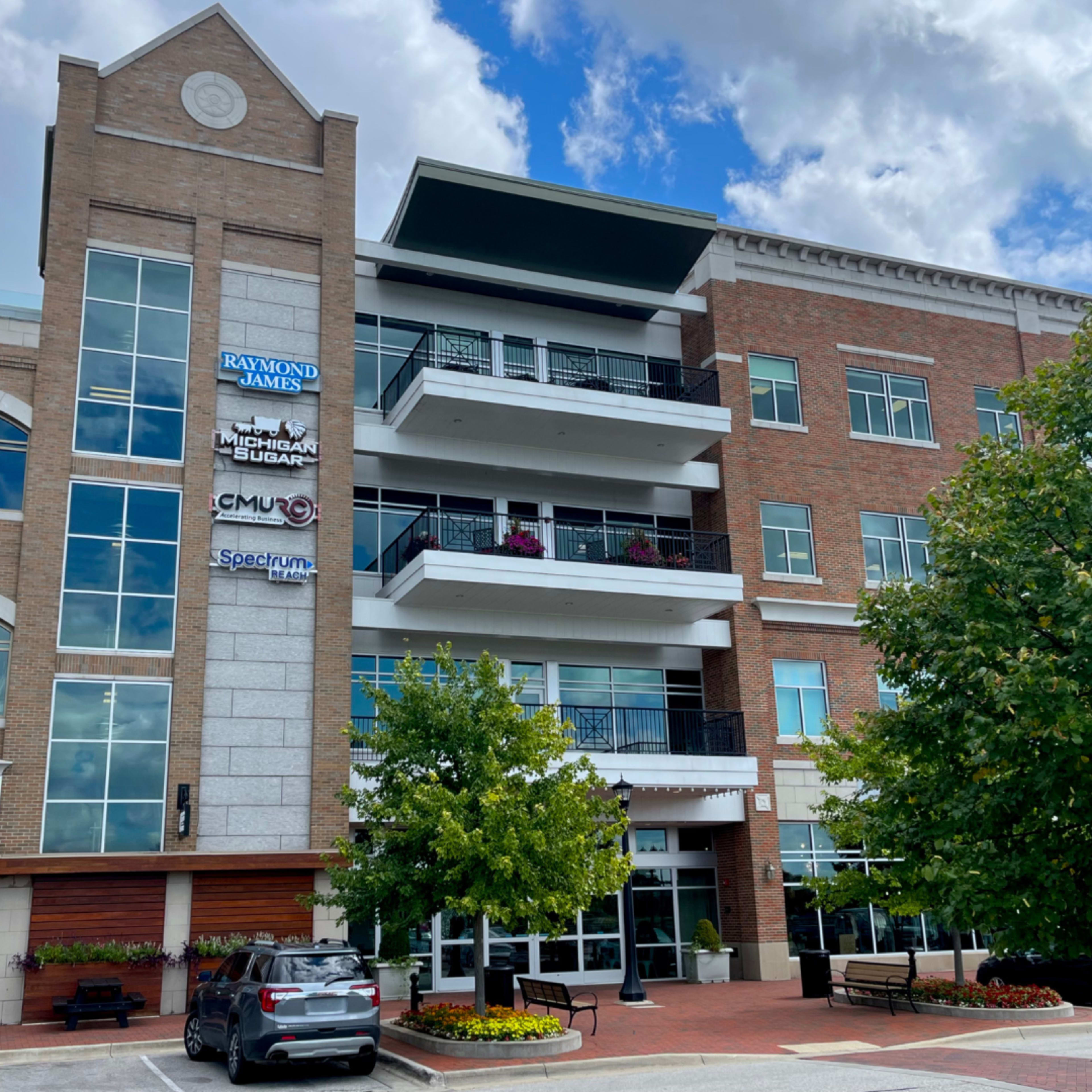 The image shows a multi-story brick building featuring several balconies, planters, and signage for various businesses, including Raymond James and Michigan Sugar.