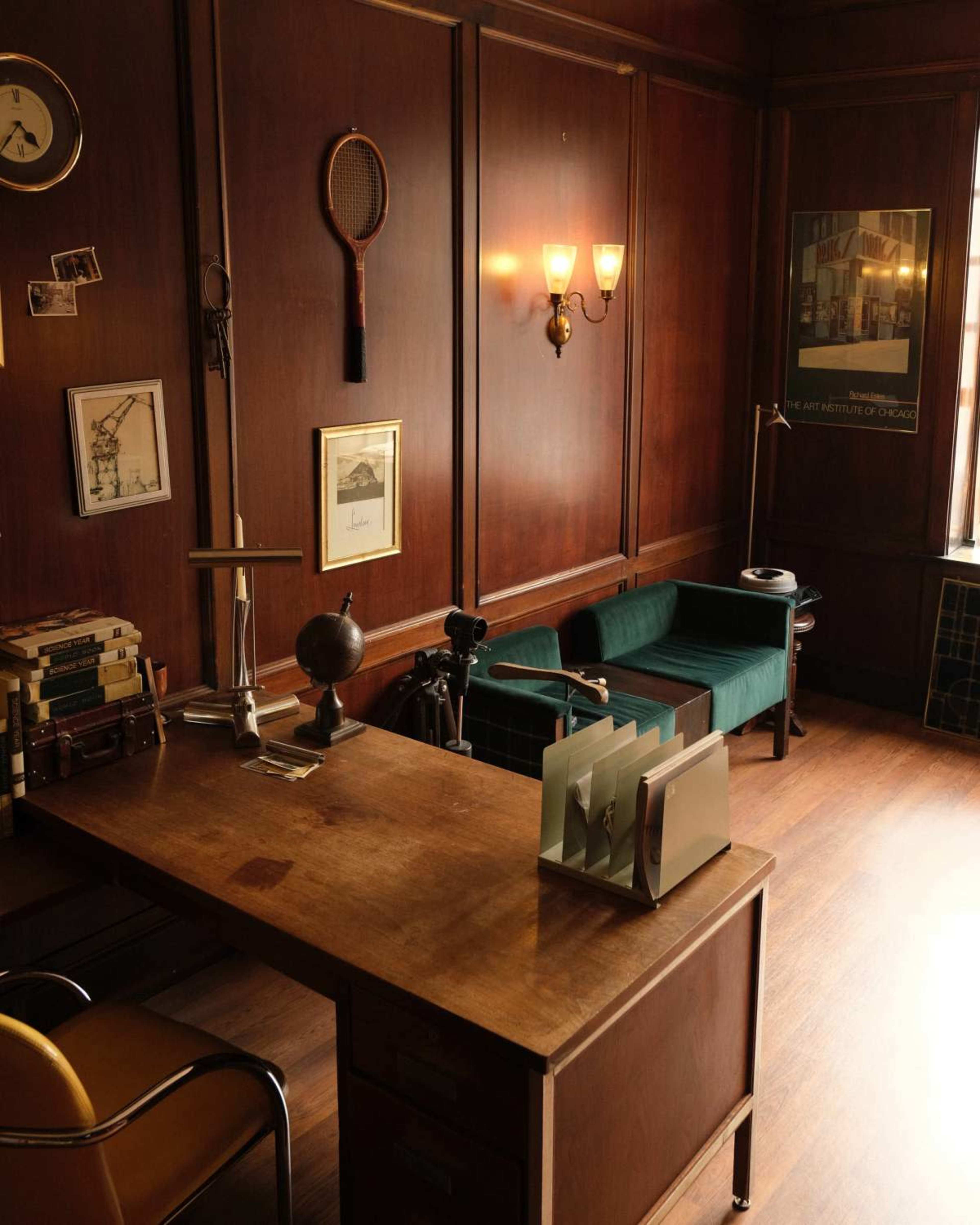The image shows a wooden office with a desk, a globe, bookshelves, and a green couch, accented by vintage decor on the walls.