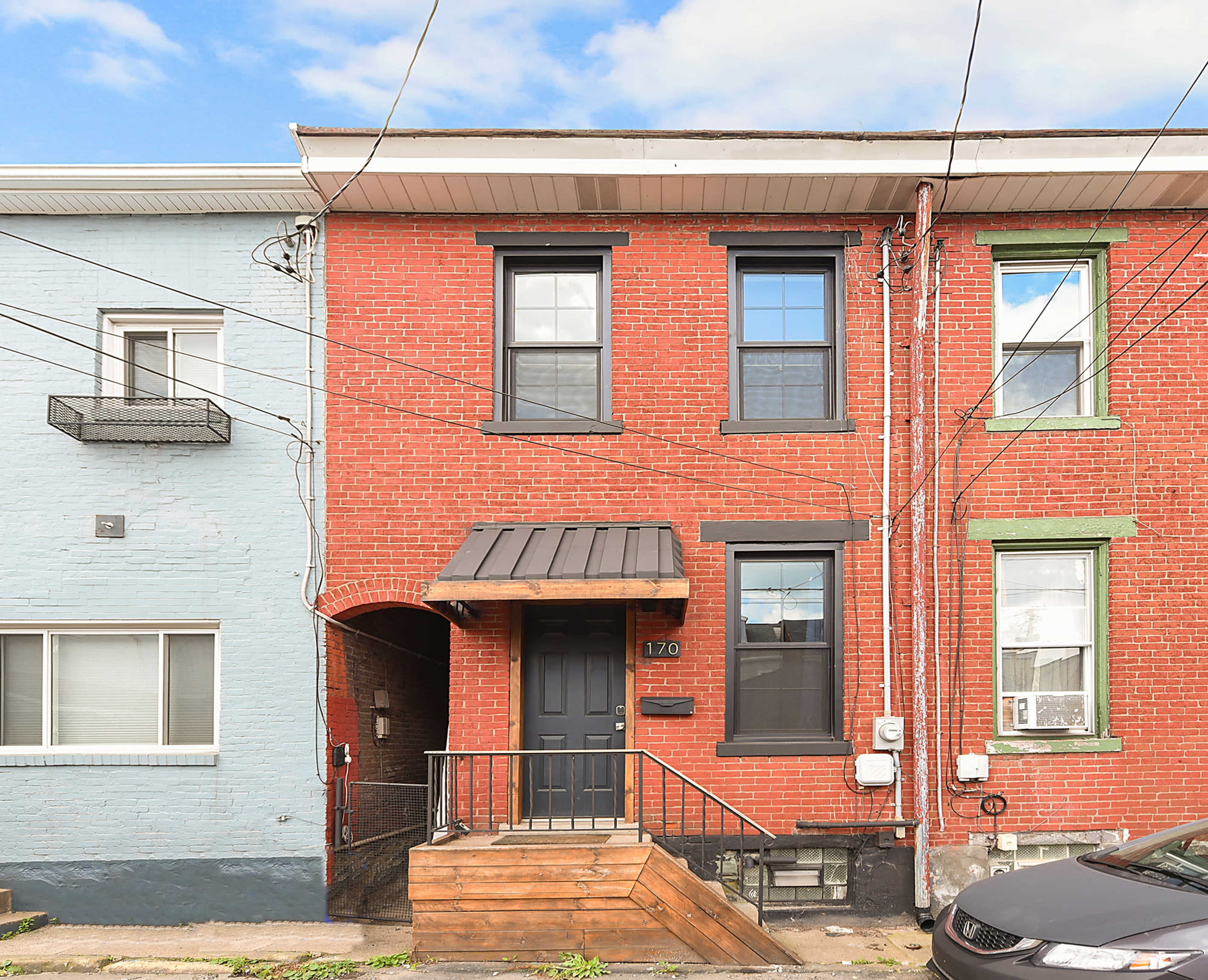 The image shows a two-story brick townhouse with a black front door and a small wooden staircase, flanked by a light blue house on the left.