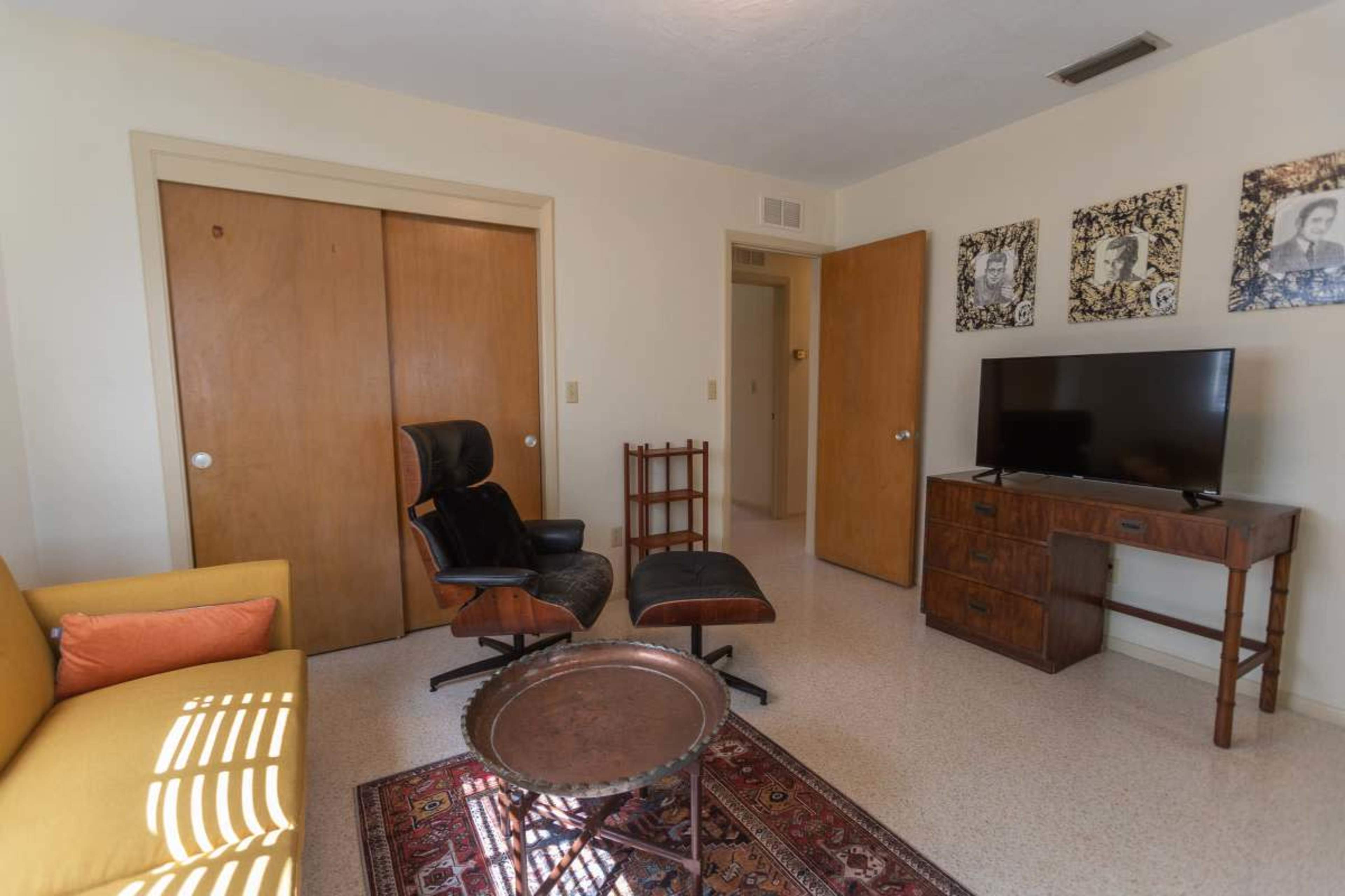 The image shows a small living room with a yellow couch, a black lounge chair, a wooden table, a television, and two wooden closets.