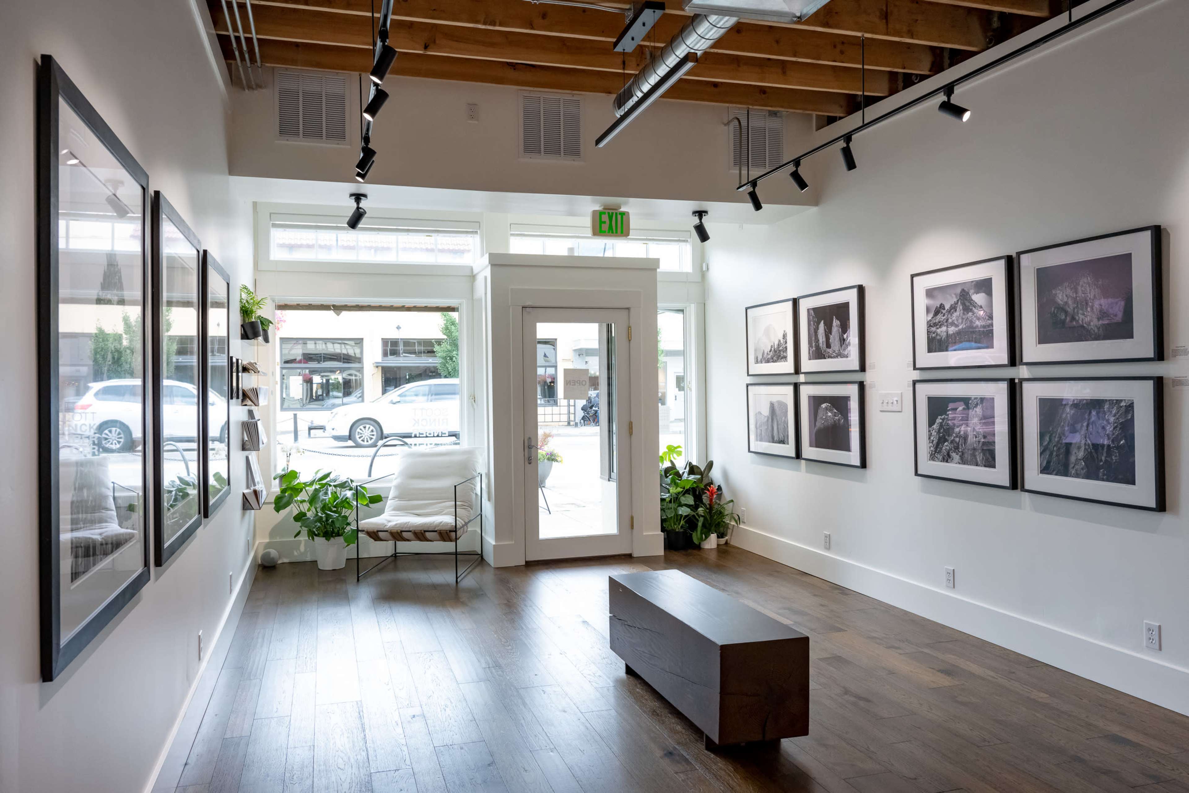 A gallery space with framed photographs on the walls, a wooden bench in the center, and large windows allowing natural light to enter.