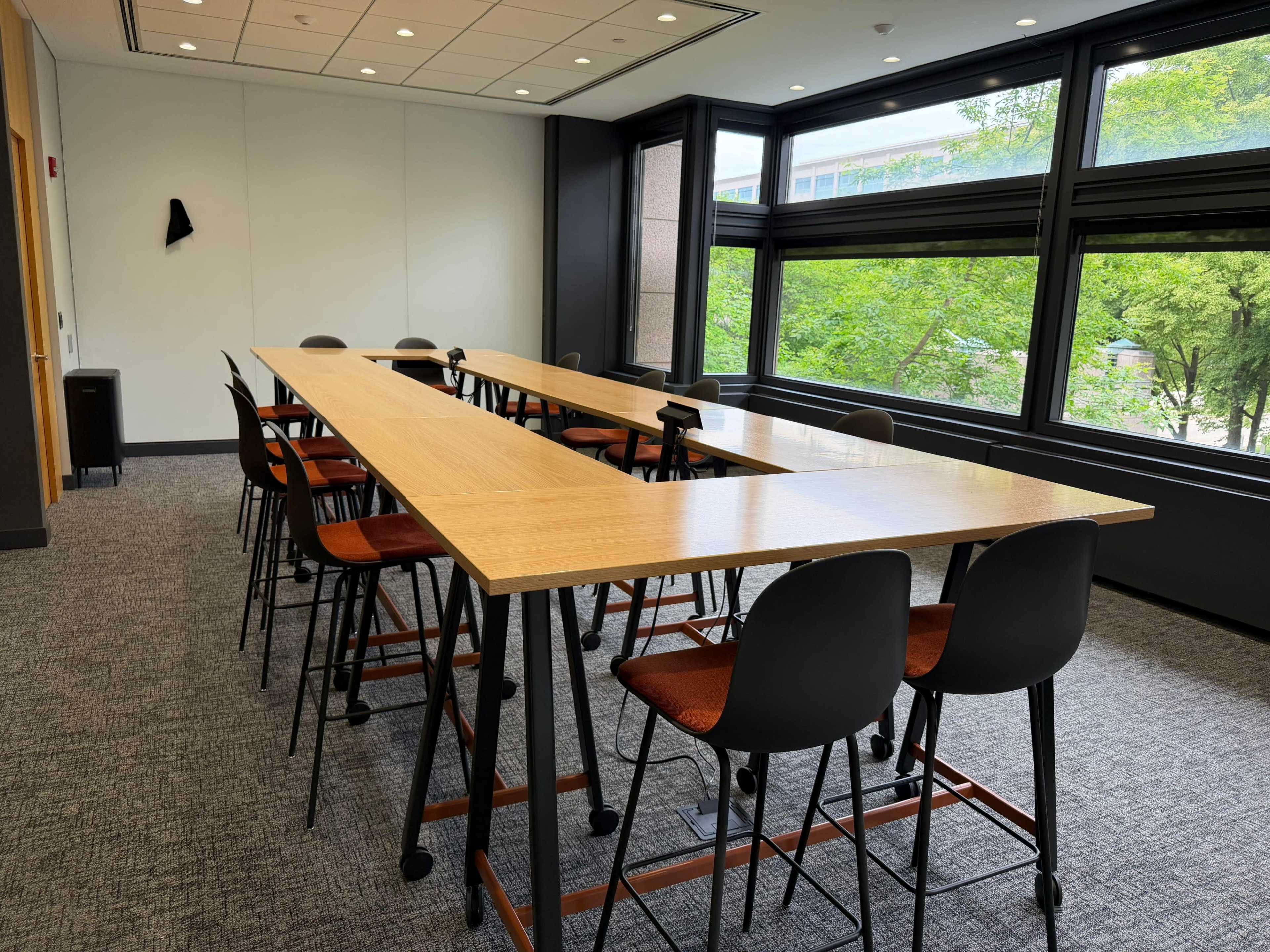 The image shows a modern meeting room with a long wooden table surrounded by high-backed chairs and large windows overlooking greenery.
