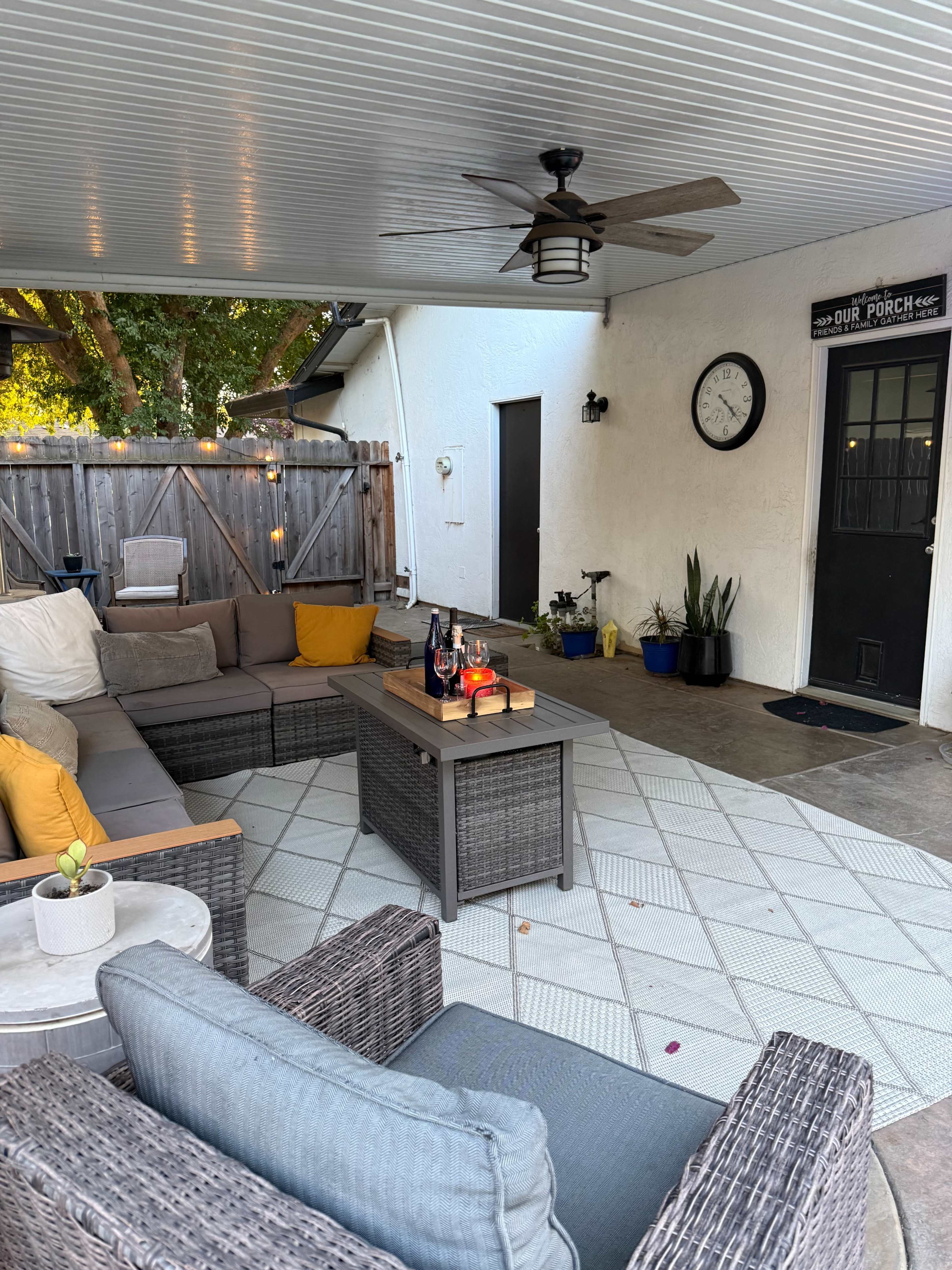 The image shows a cozy outdoor porch area furnished with a sectional sofa, a coffee table, and a wall clock, surrounded by greenery and a wooden fence.