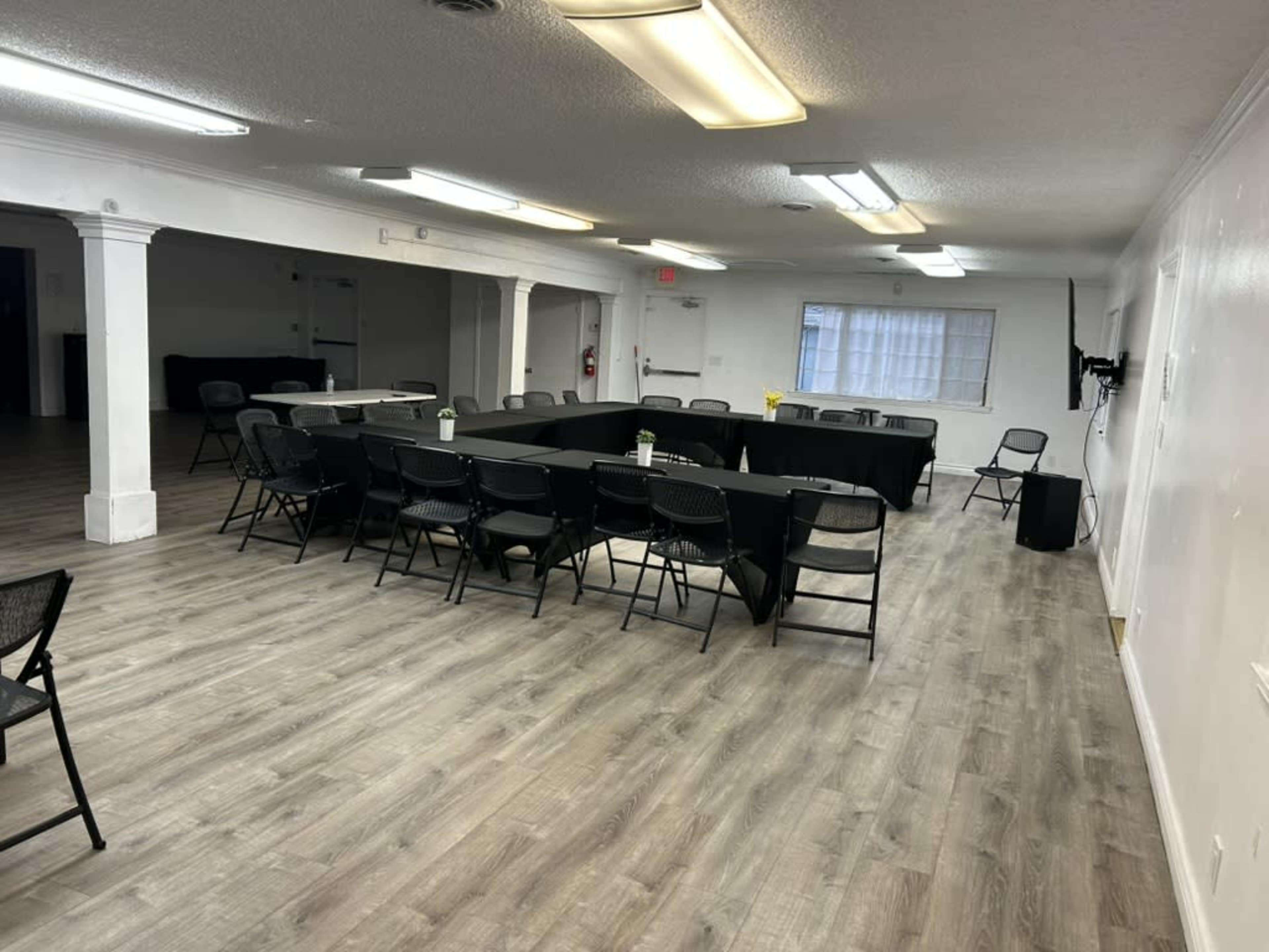 The image shows a spacious meeting room set up with a large rectangular table surrounded by folding chairs, with a polished wooden floor and overhead lighting.