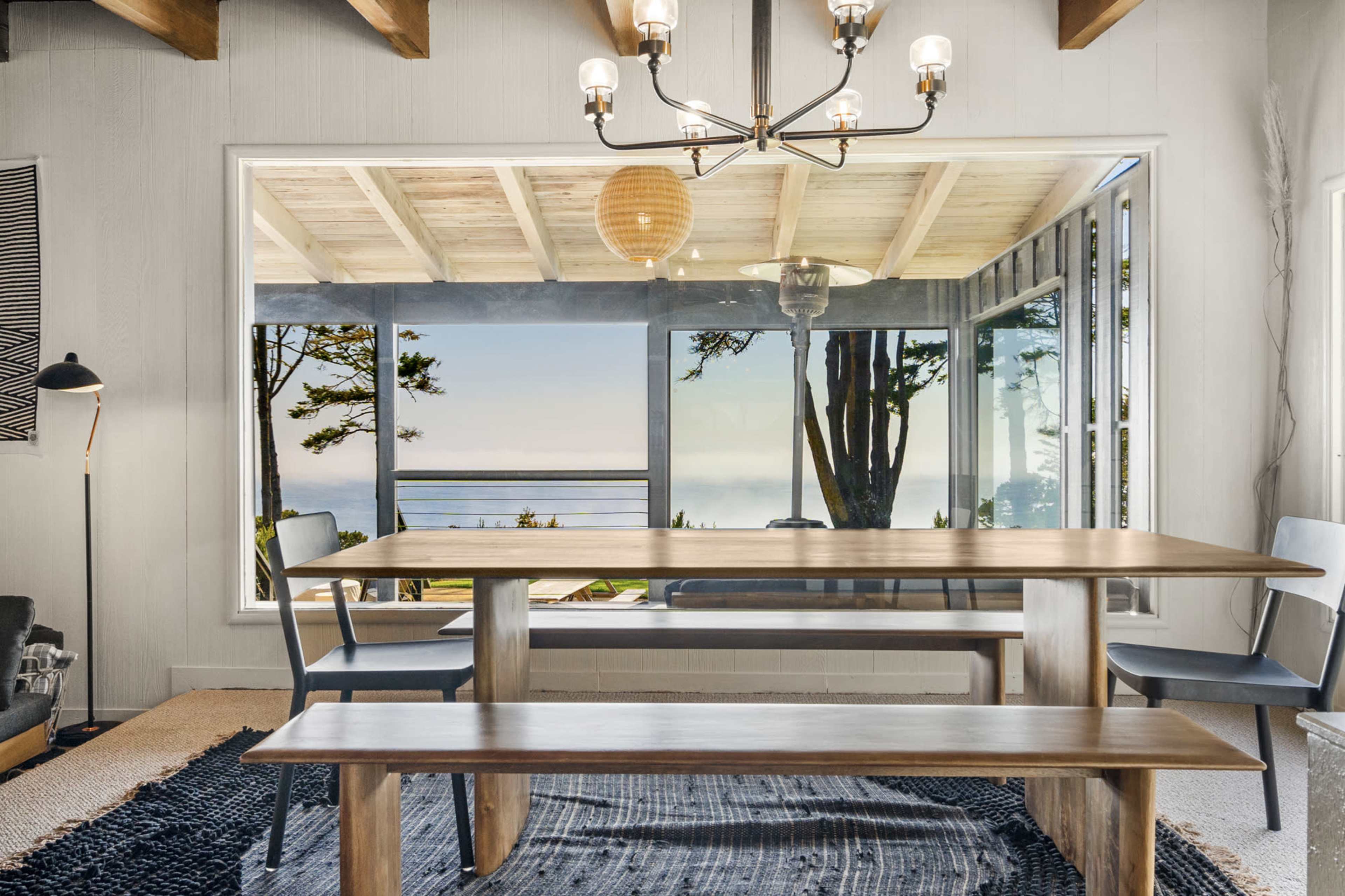 A dining area features a wooden table and benches with large windows overlooking a scenic view of the ocean and trees.
