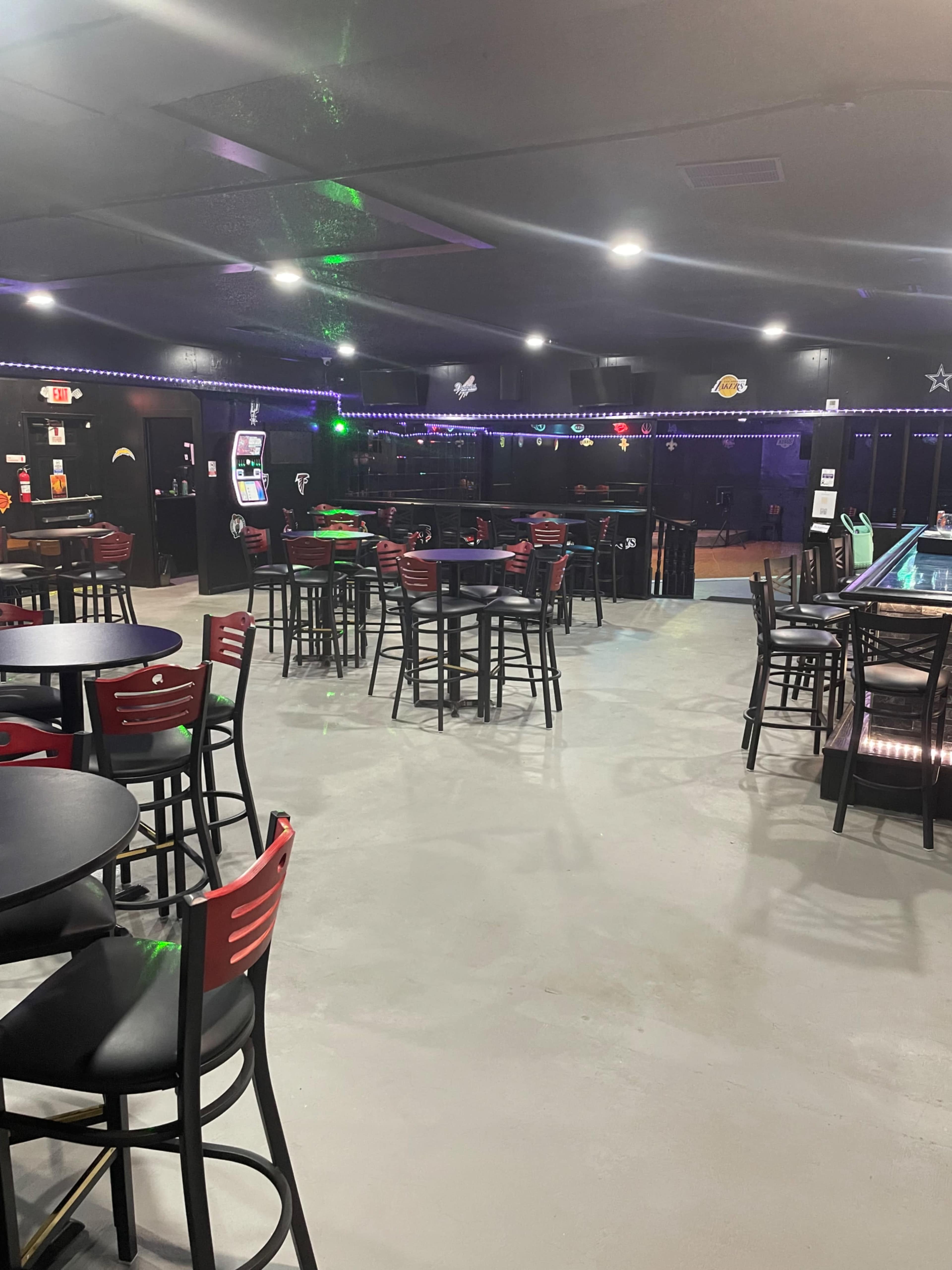 The image shows an empty bar area with high tables and chairs, neon lights, and various gaming machines in the background.
