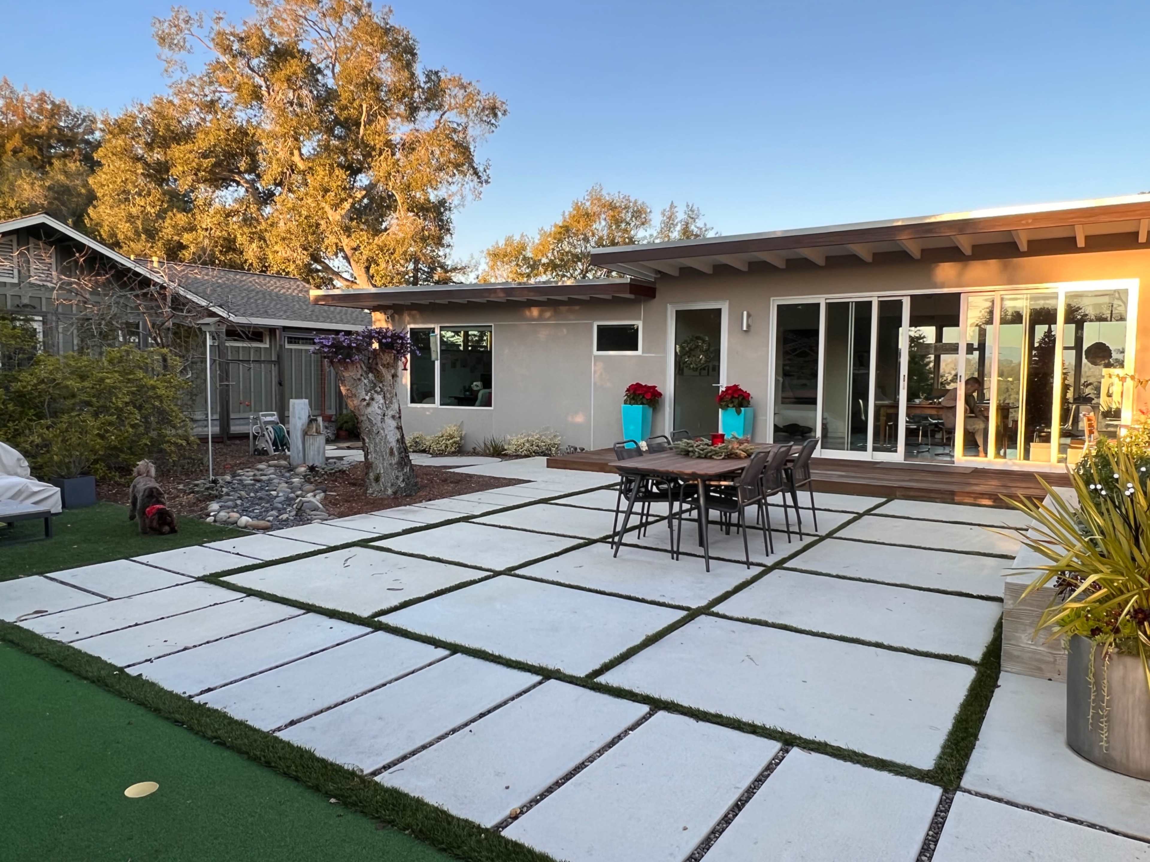 A modern backyard patio featuring a large concrete paver area, a seating arrangement with chairs, and decorative plants in planters.
