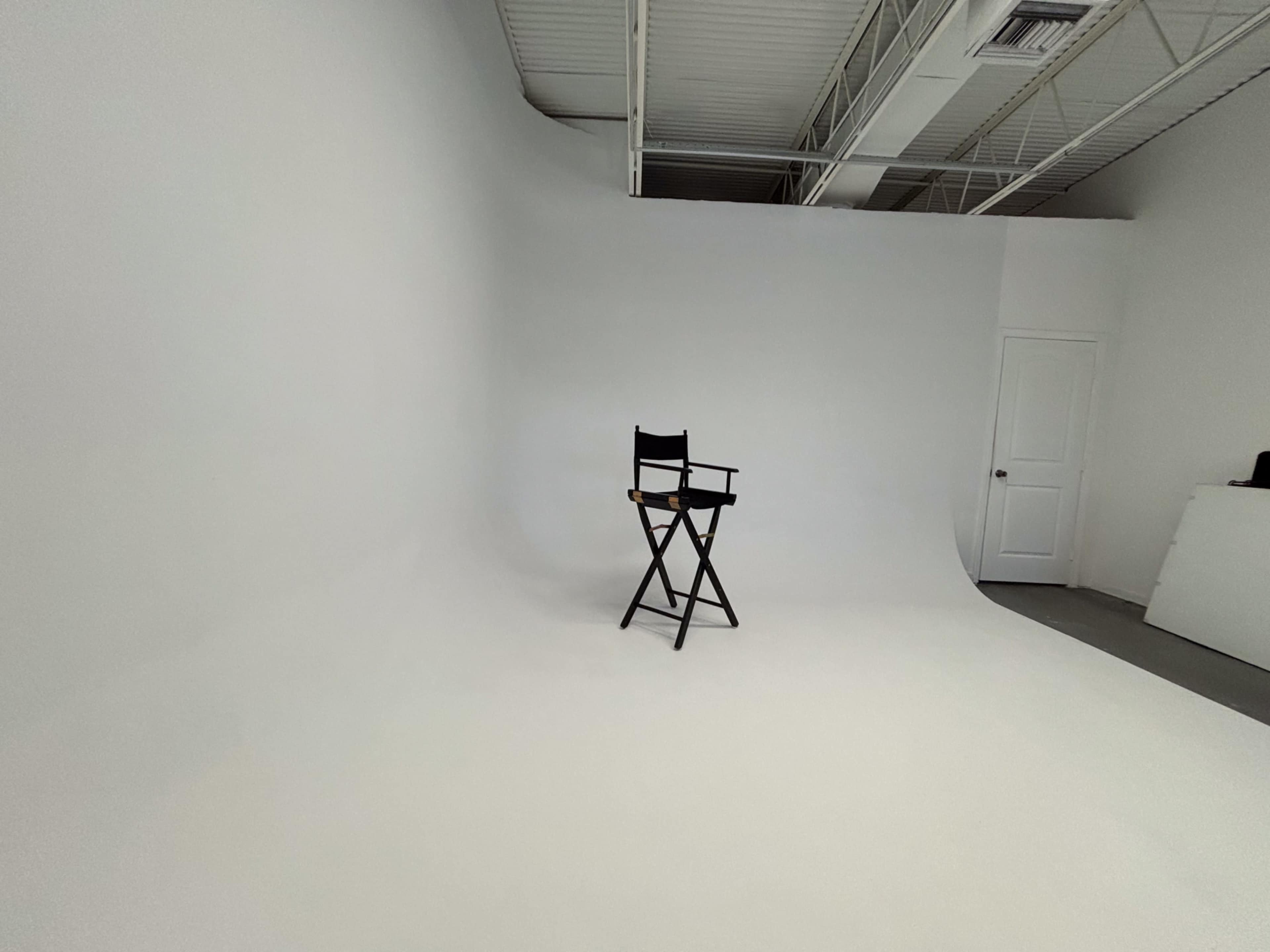 A black director's chair stands alone in a spacious, empty photo studio with a seamless white backdrop.
