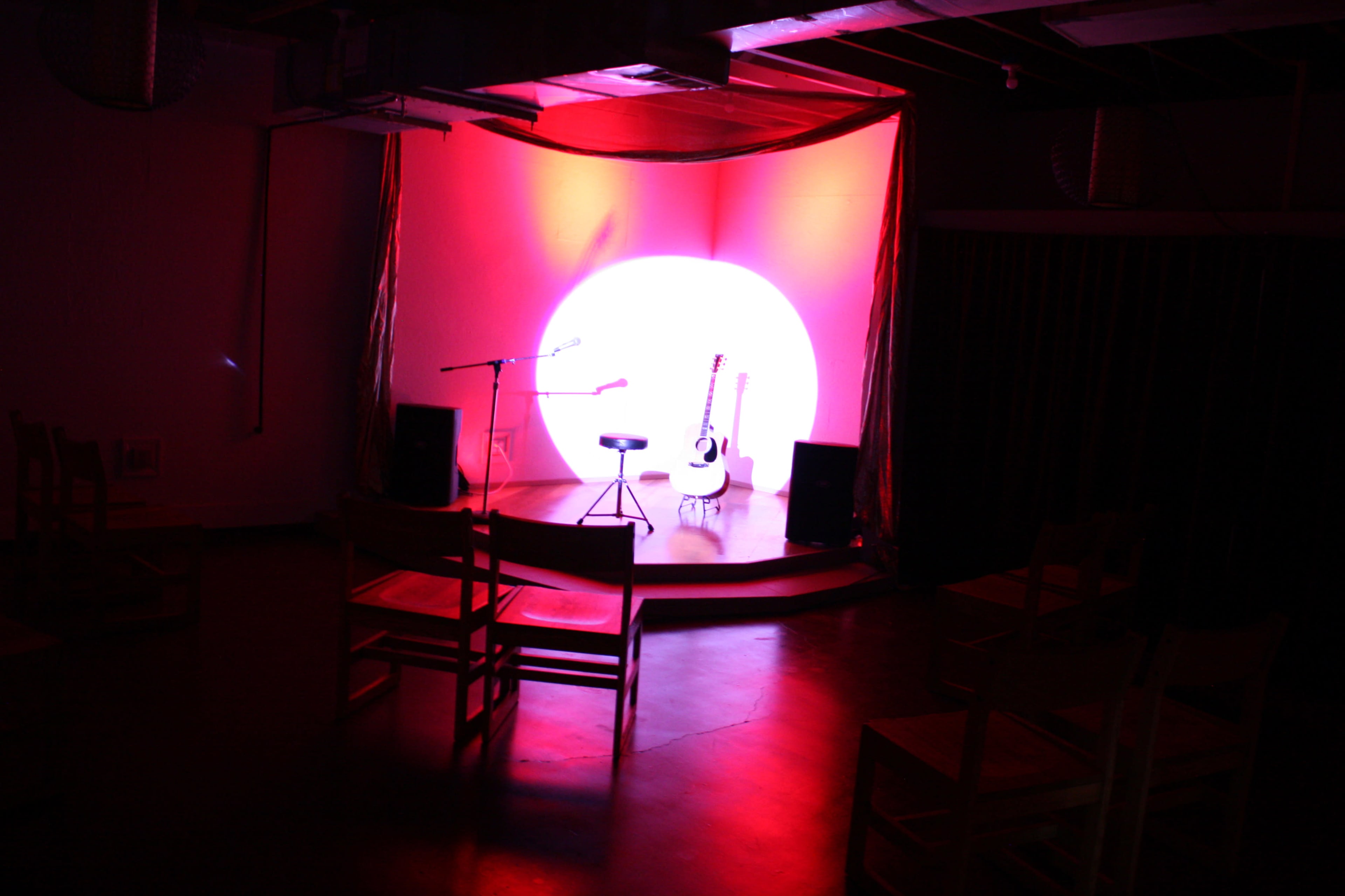 A spotlight illuminates an empty stage with a microphone, a stool, and an acoustic guitar in front of a large circular backdrop.