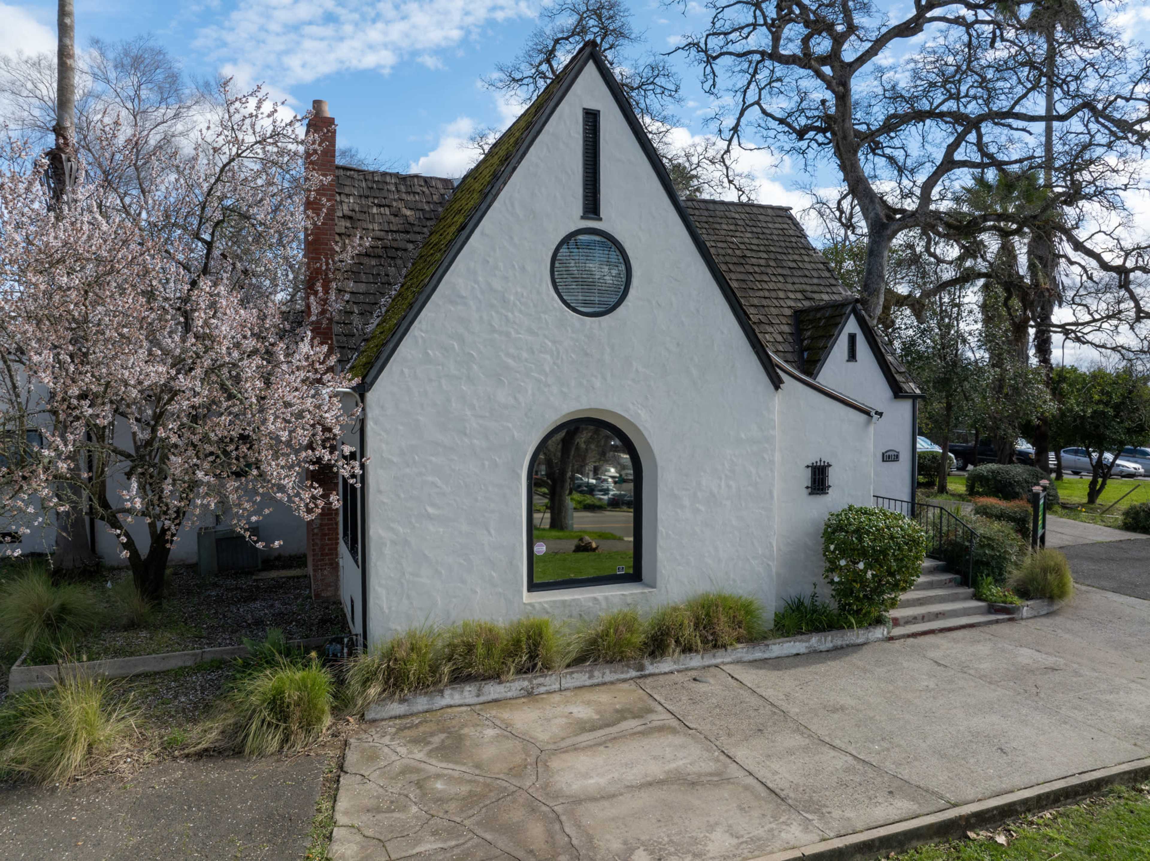 A whitewashed house with a peaked roof and a large front window is flanked by flowering trees and greenery.