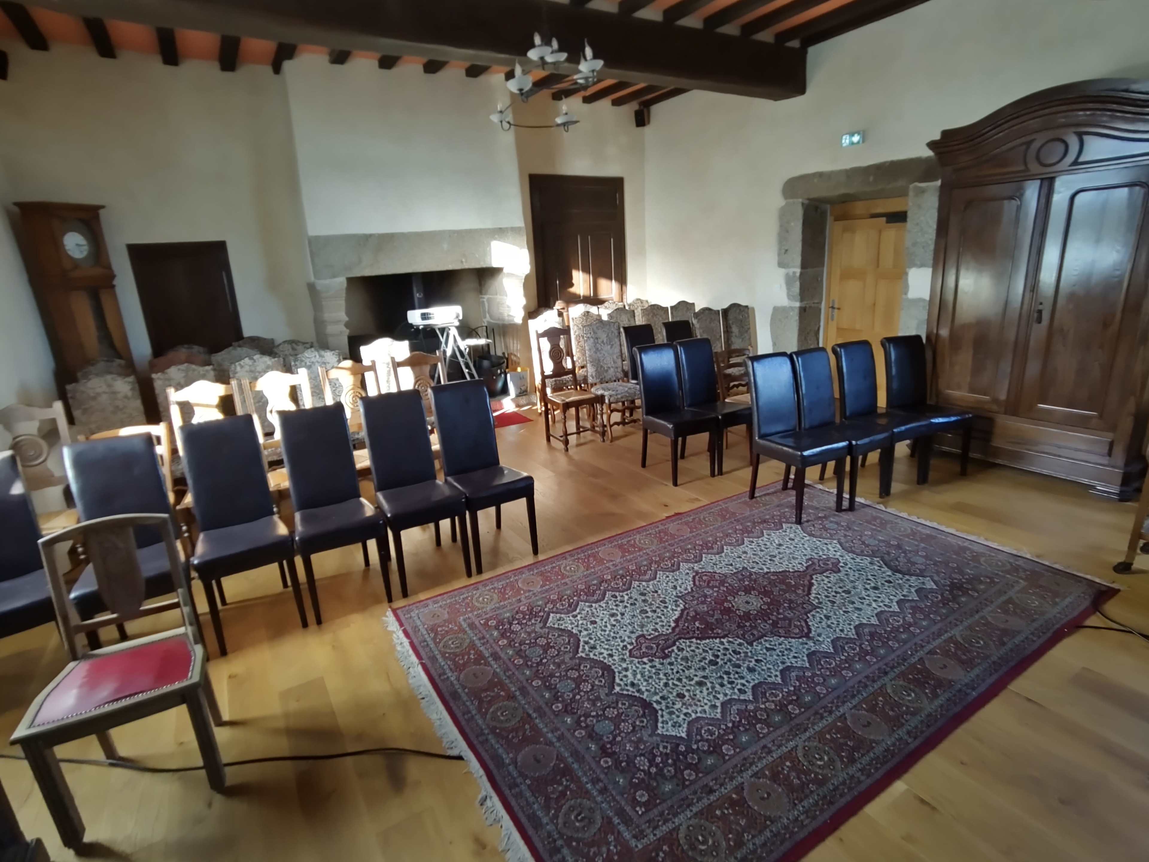 The image shows a spacious room with wooden floors, featuring rows of chairs arranged toward a large area rug, a wooden wardrobe, and a clock on the wall.