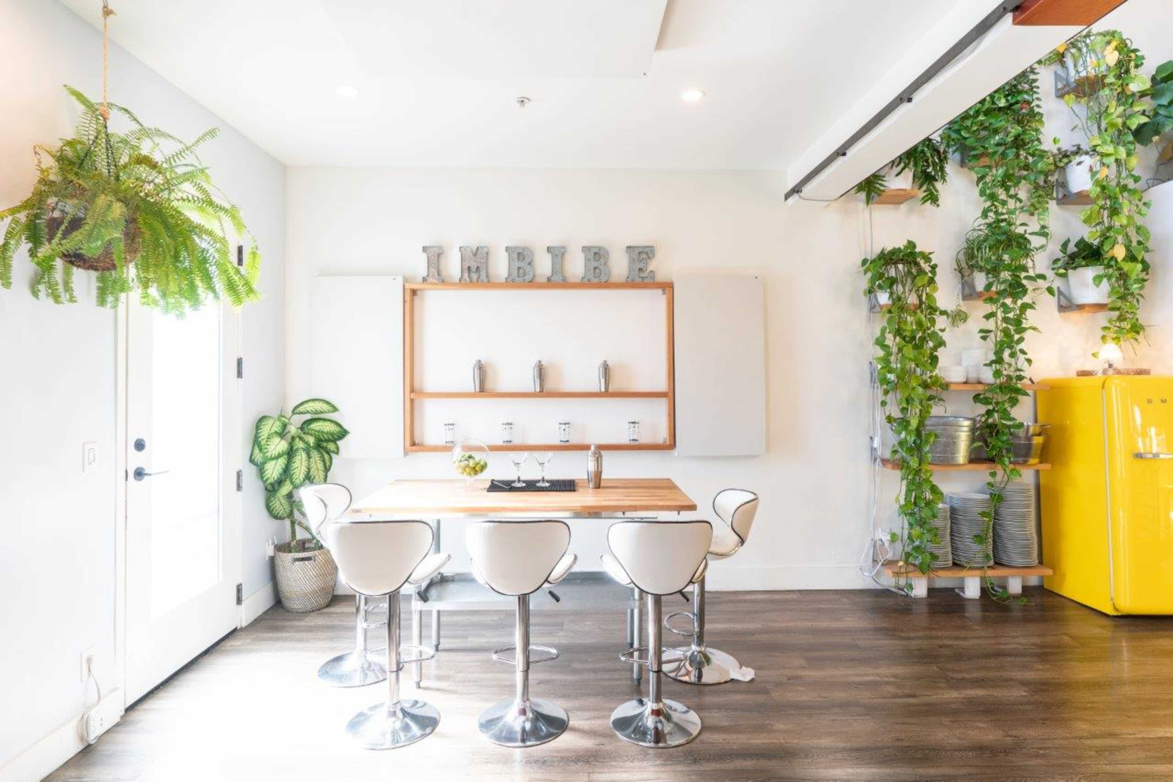 A modern dining area features a wooden table surrounded by white stools, with green plants hanging and a yellow refrigerator in the background.