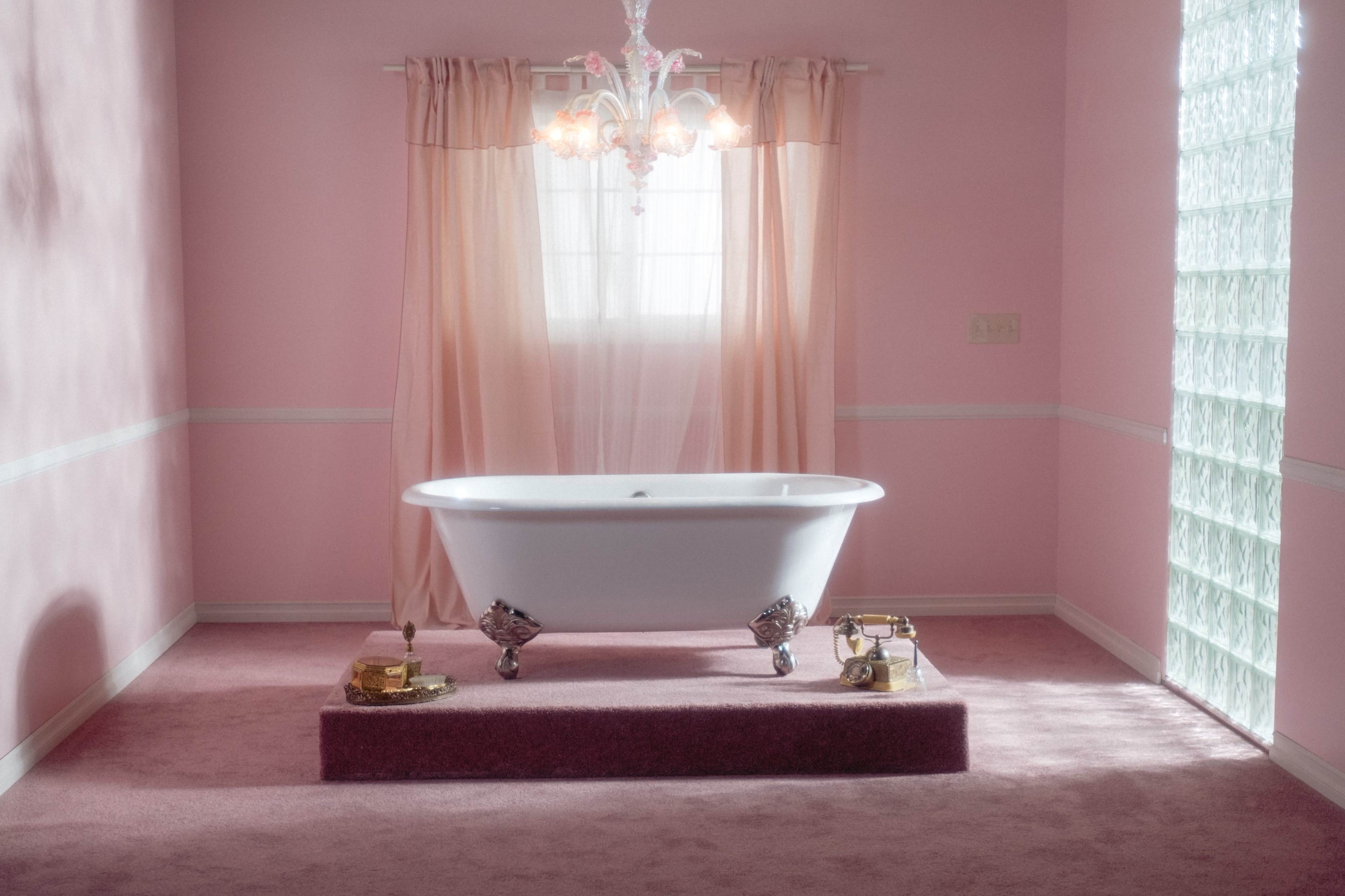 A white clawfoot bathtub sits on a raised platform in a pink bathroom, with a chandelier overhead and light filtering through sheer curtains.