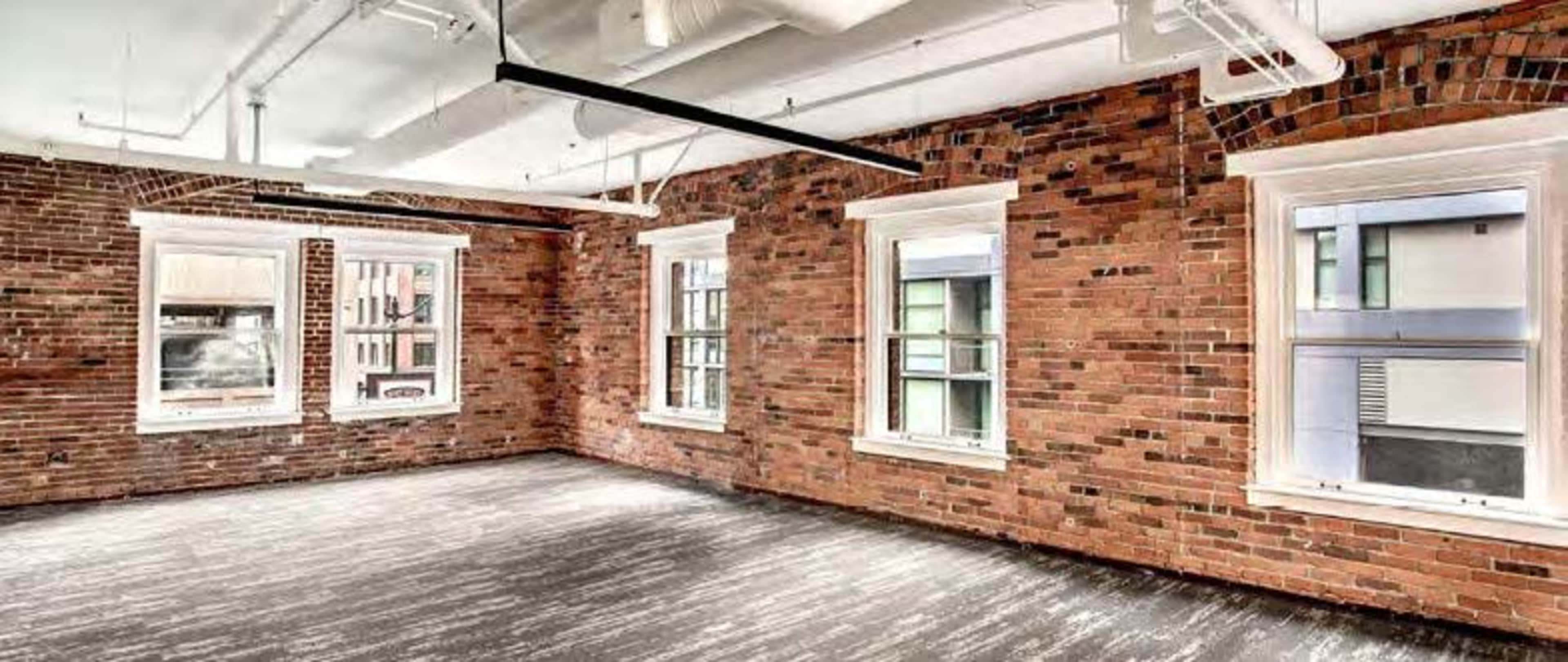 The image shows an empty room with exposed brick walls and four windows allowing natural light.