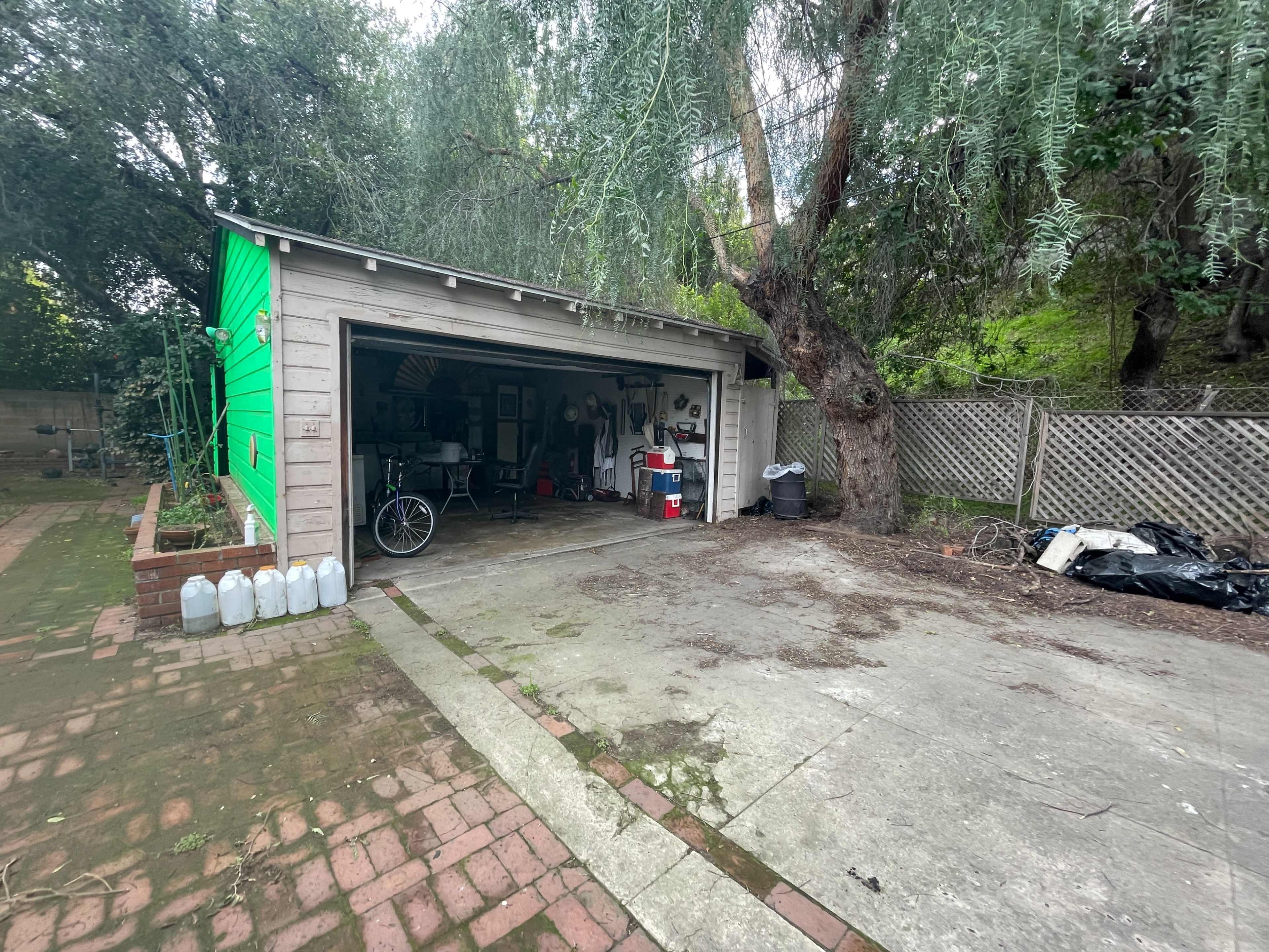 A garage with a partially open door reveals various tools and items inside, while a tree looms nearby in a cluttered outdoor area.