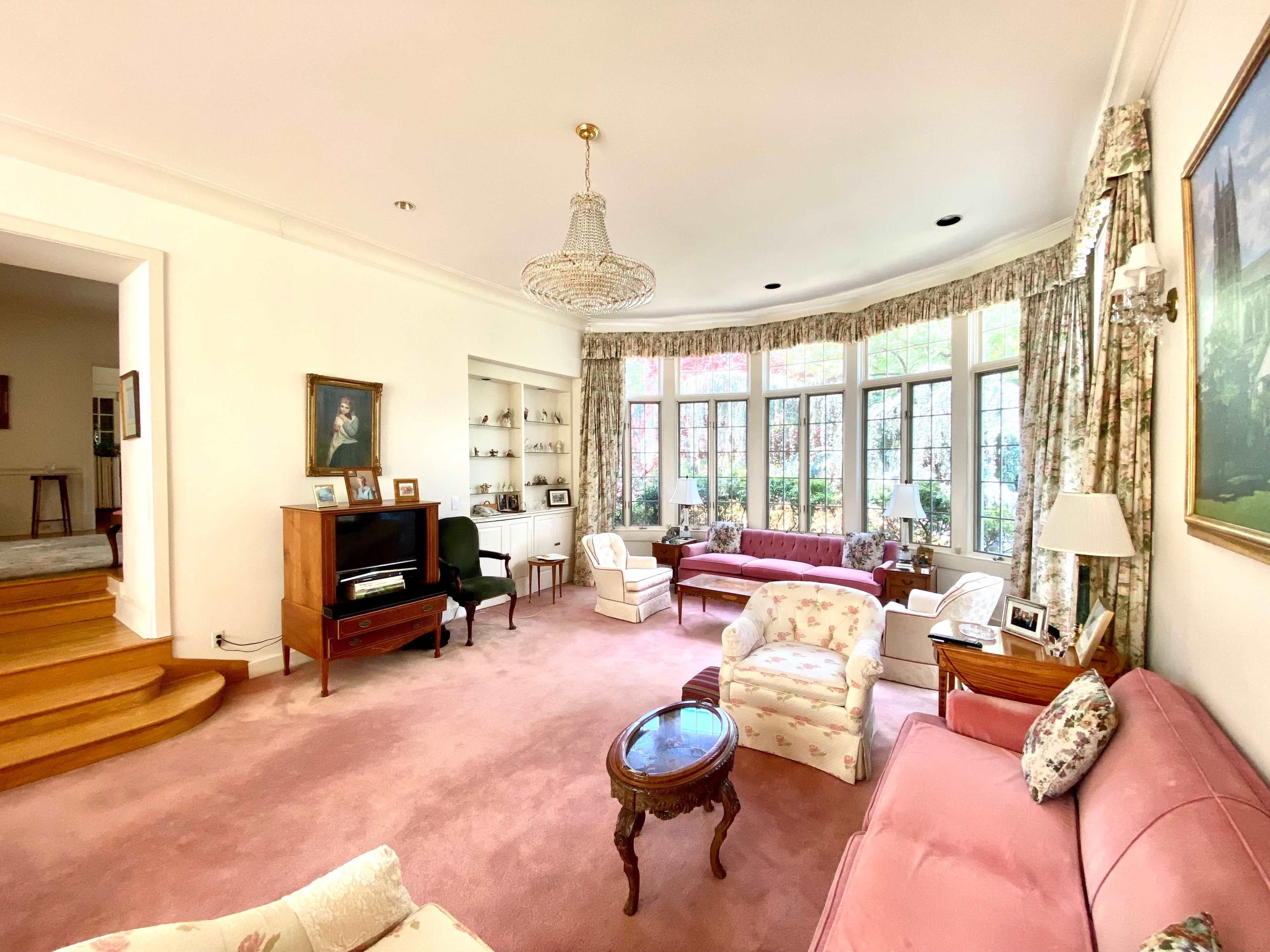The living room features a pink carpet, floral-patterned furniture, large windows with views of greenery, and a chandelier.