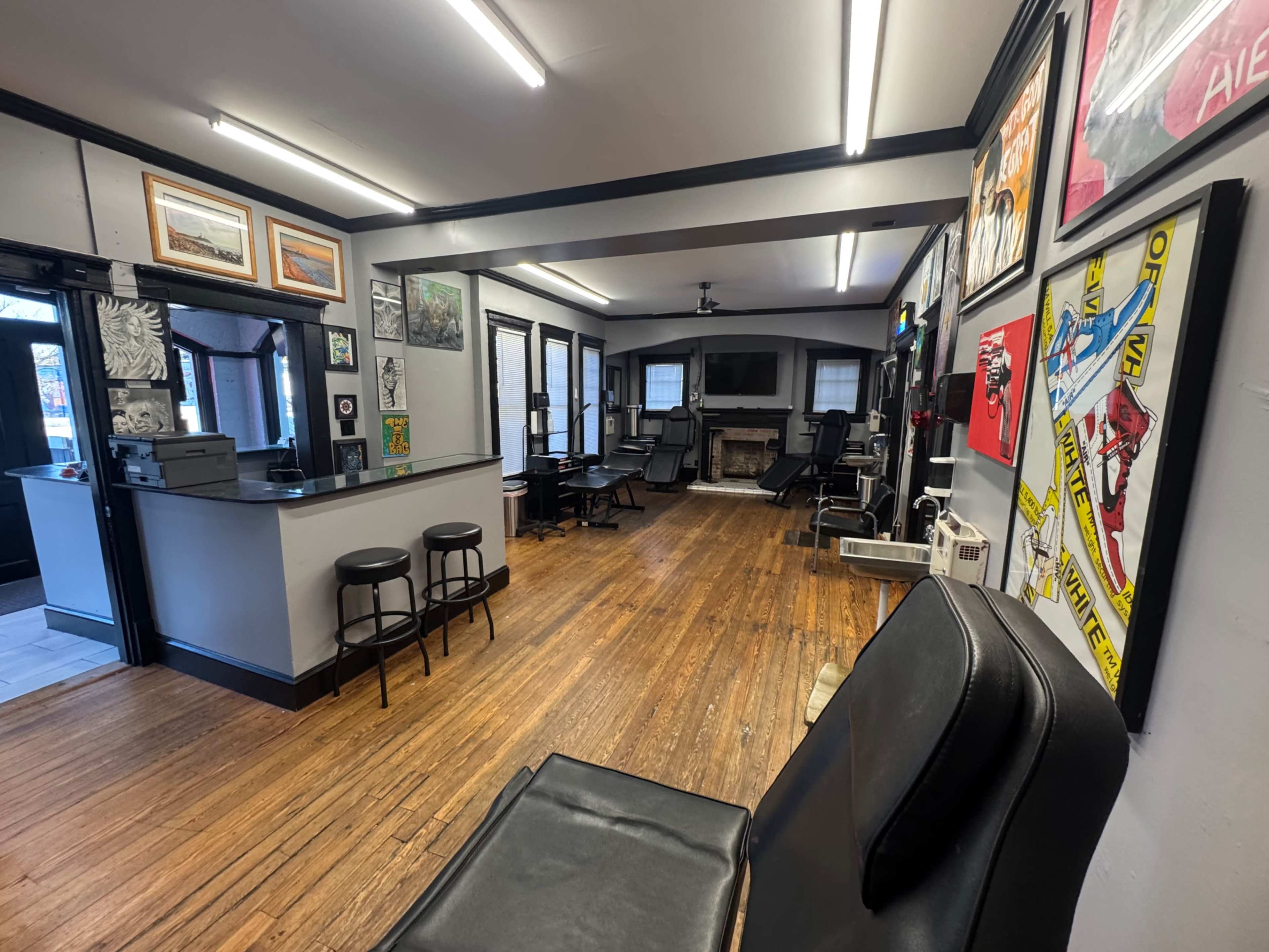 The image shows a spacious room with wooden floors, featuring several art pieces on the walls, a reception area with barstools, and various seating arrangements.