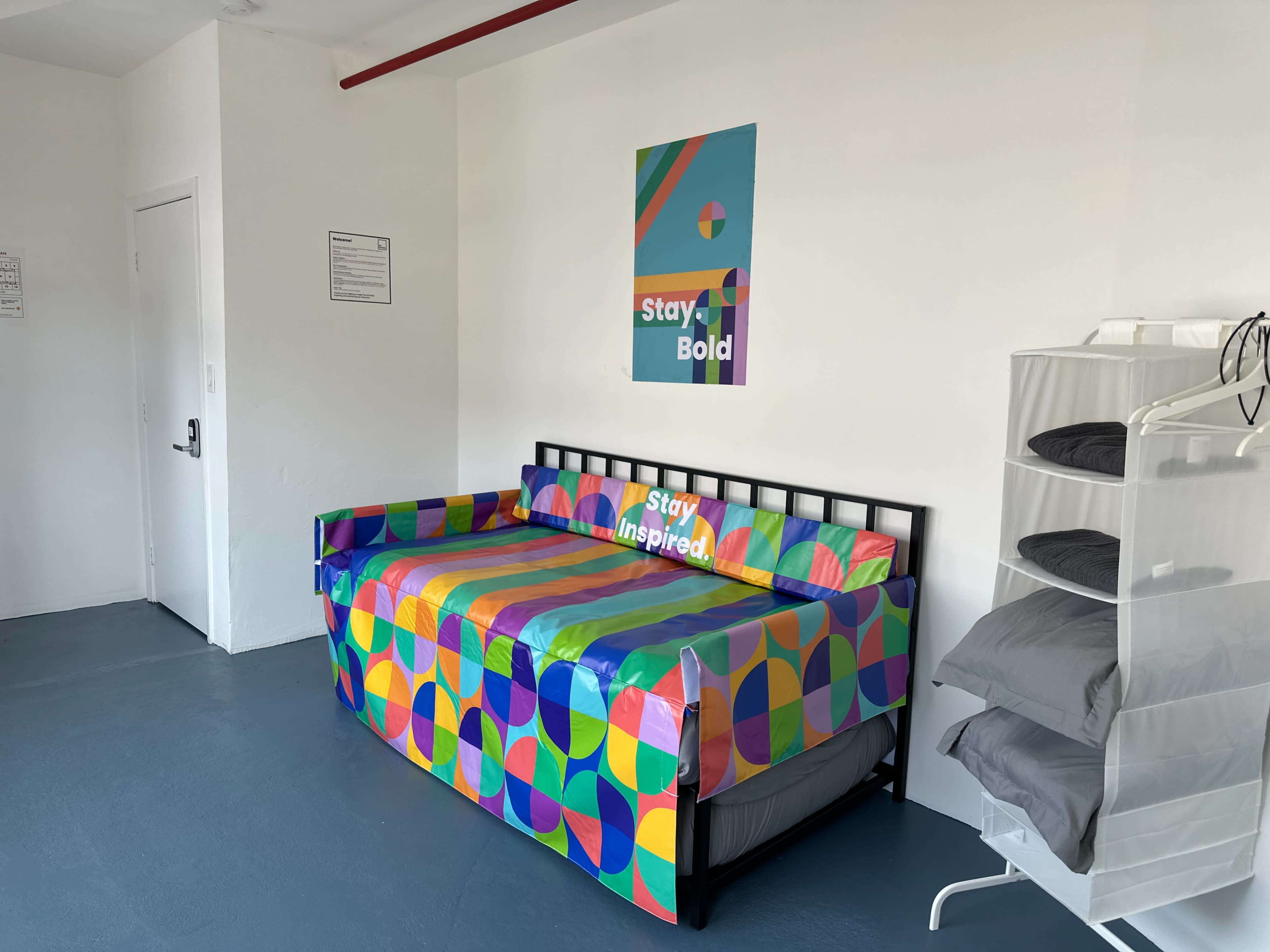 The image shows a colorful daybed with a geometric-patterned cover, positioned against a white wall featuring a motivational poster that reads "Stay Bold" and "Stay Inspired."