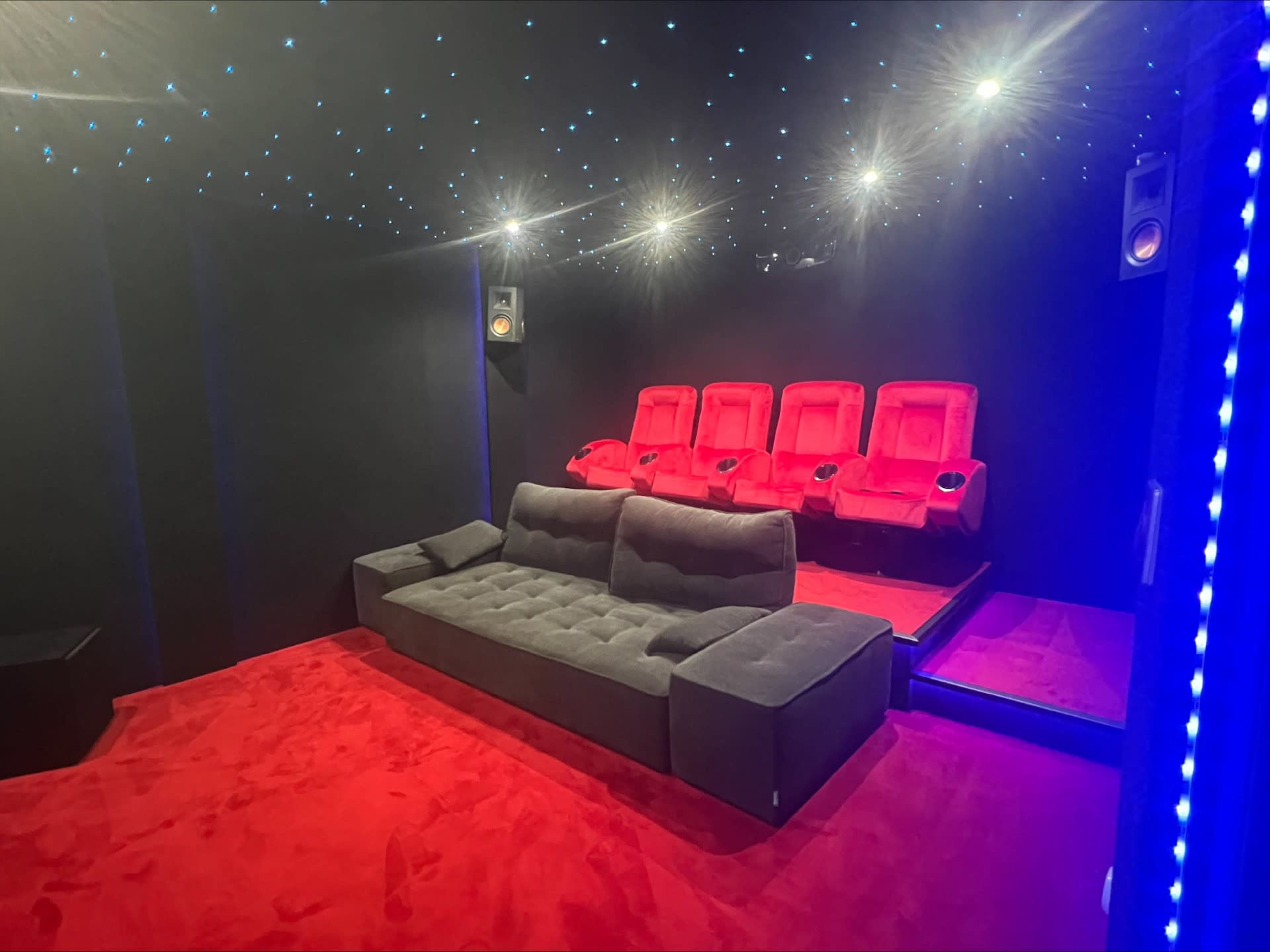 The image shows a dimly lit home theater featuring a gray sofa and red recliner seats arranged against a black wall with a starry ceiling.