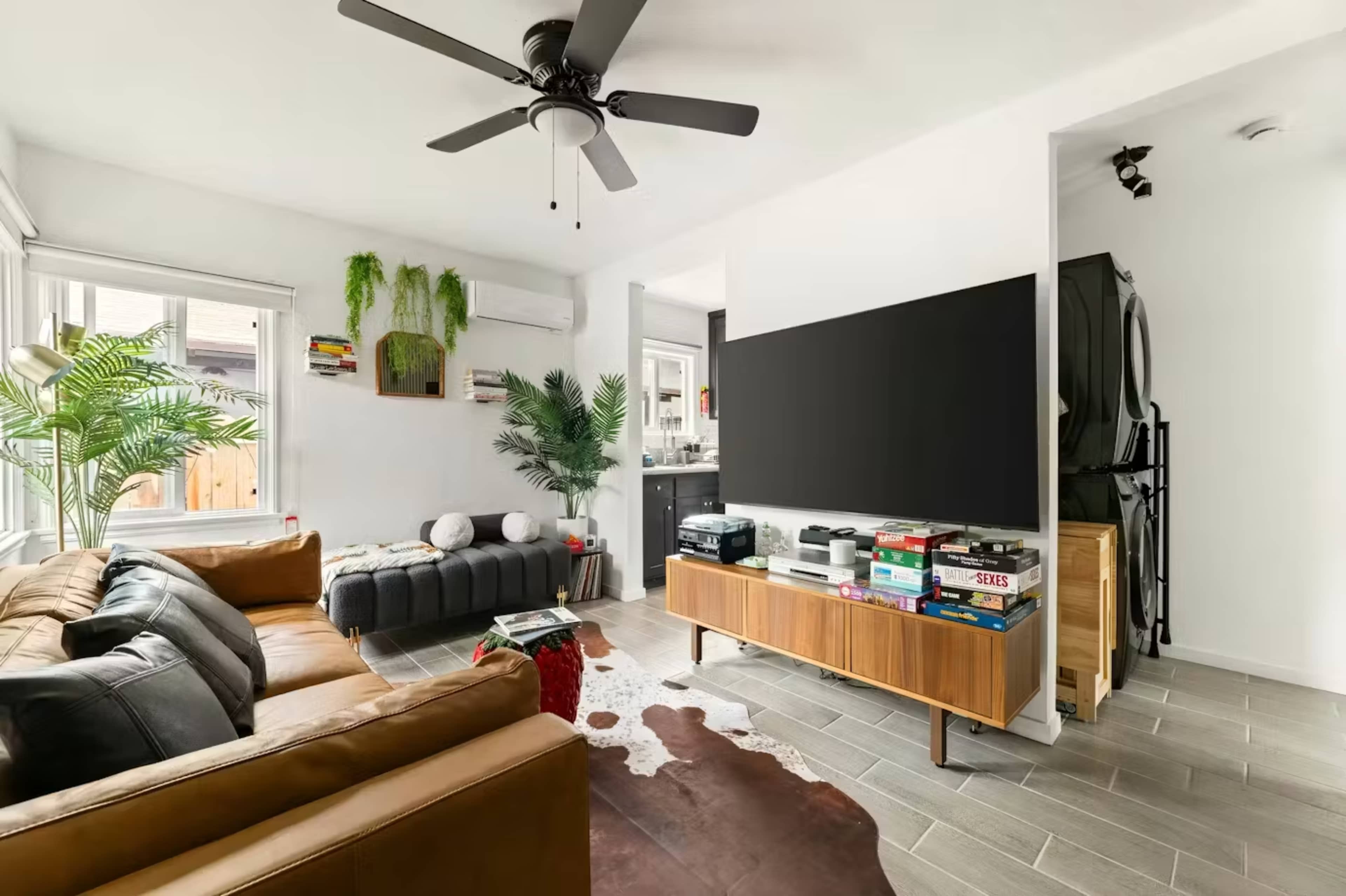A modern living room features a brown leather sofa, a large television on a wooden console, and plants alongside a stylish rug.
